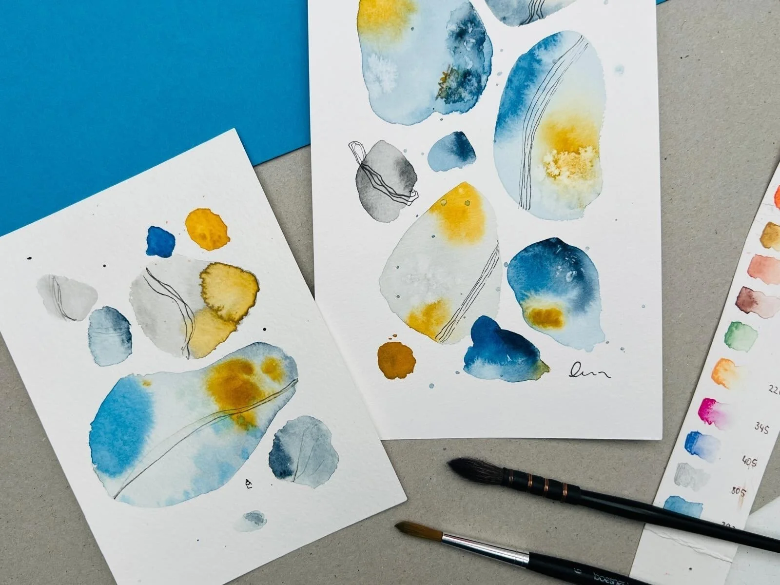 Zwei Papiere mit abstrakten Aquarellmalerei in Blau- und Gelbtönen, daneben 2 Aquarellpinsel