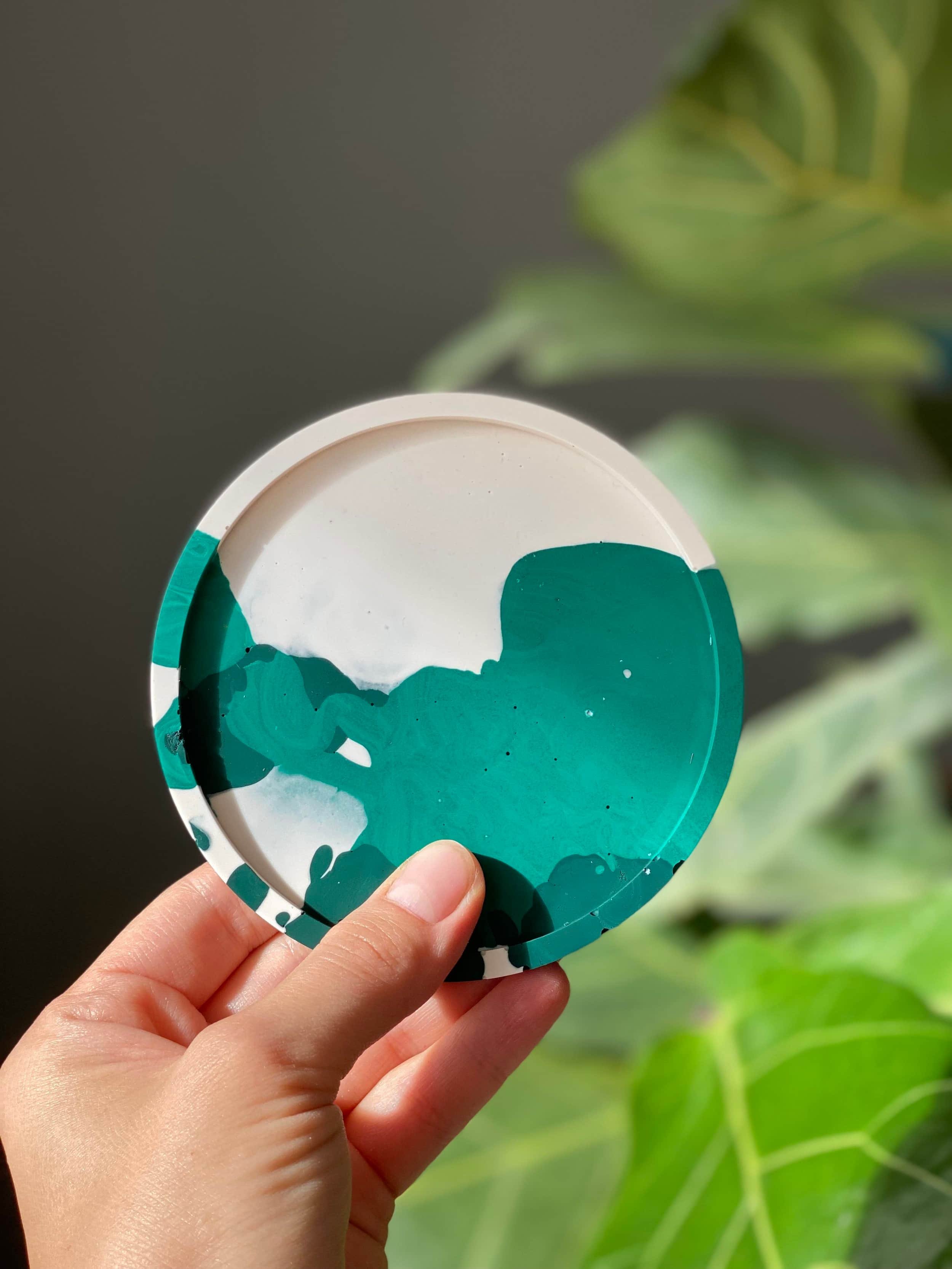 Jesmonite Untersetzer in weiß und grün, gehalten in einer Hand vor einer grünen Pflanzenwand