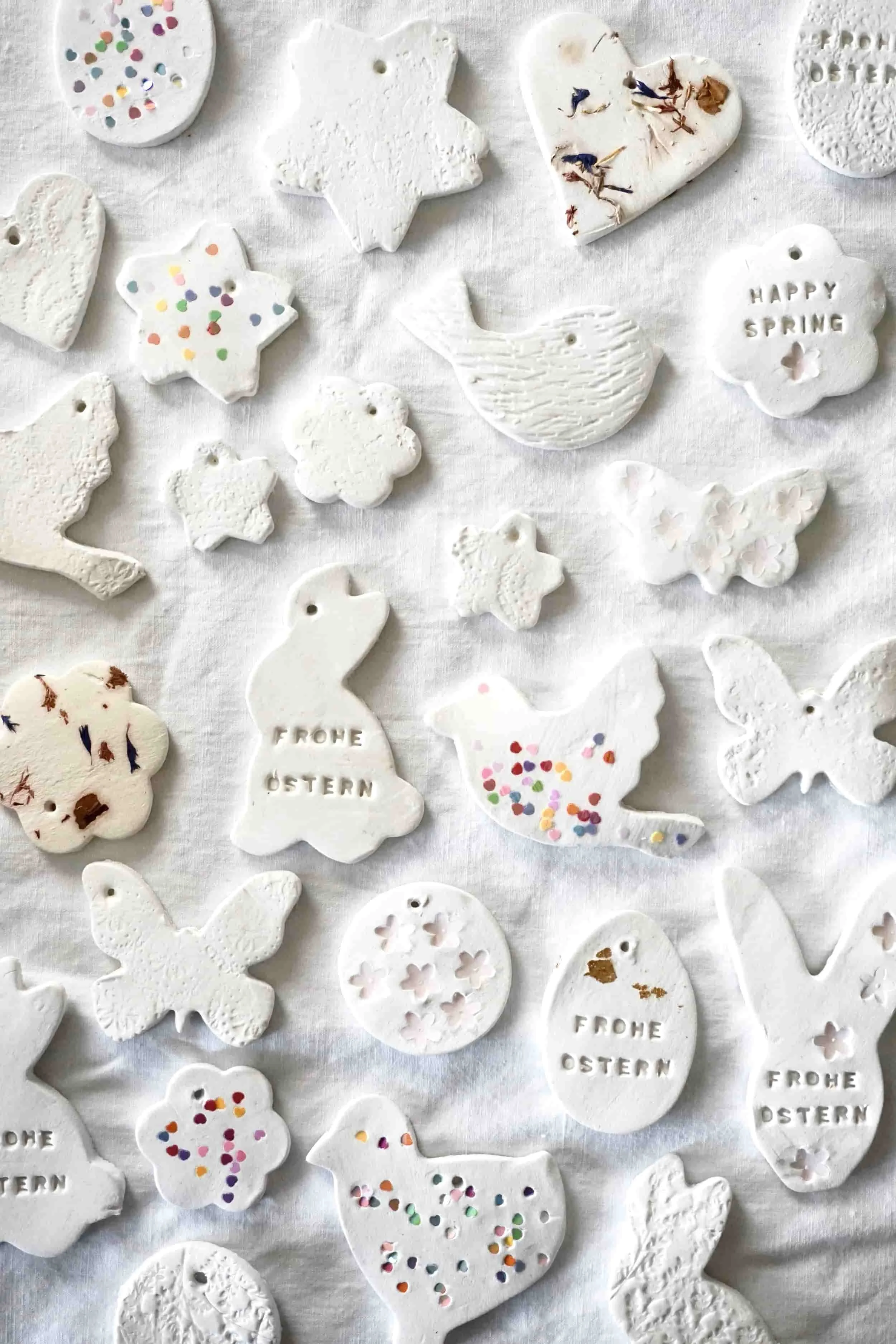 Weiße Osteranhänger in verschiedenen Formen wie Hühnern, Eiern, Schmetterlingen, Tauben, Wolken und Sternen, einige mit bunten Konfetti, andere mit eingravierten Grüßen wie 'Frohe Ostern' und 'Happy Spring', liegen auf weißem Stoff