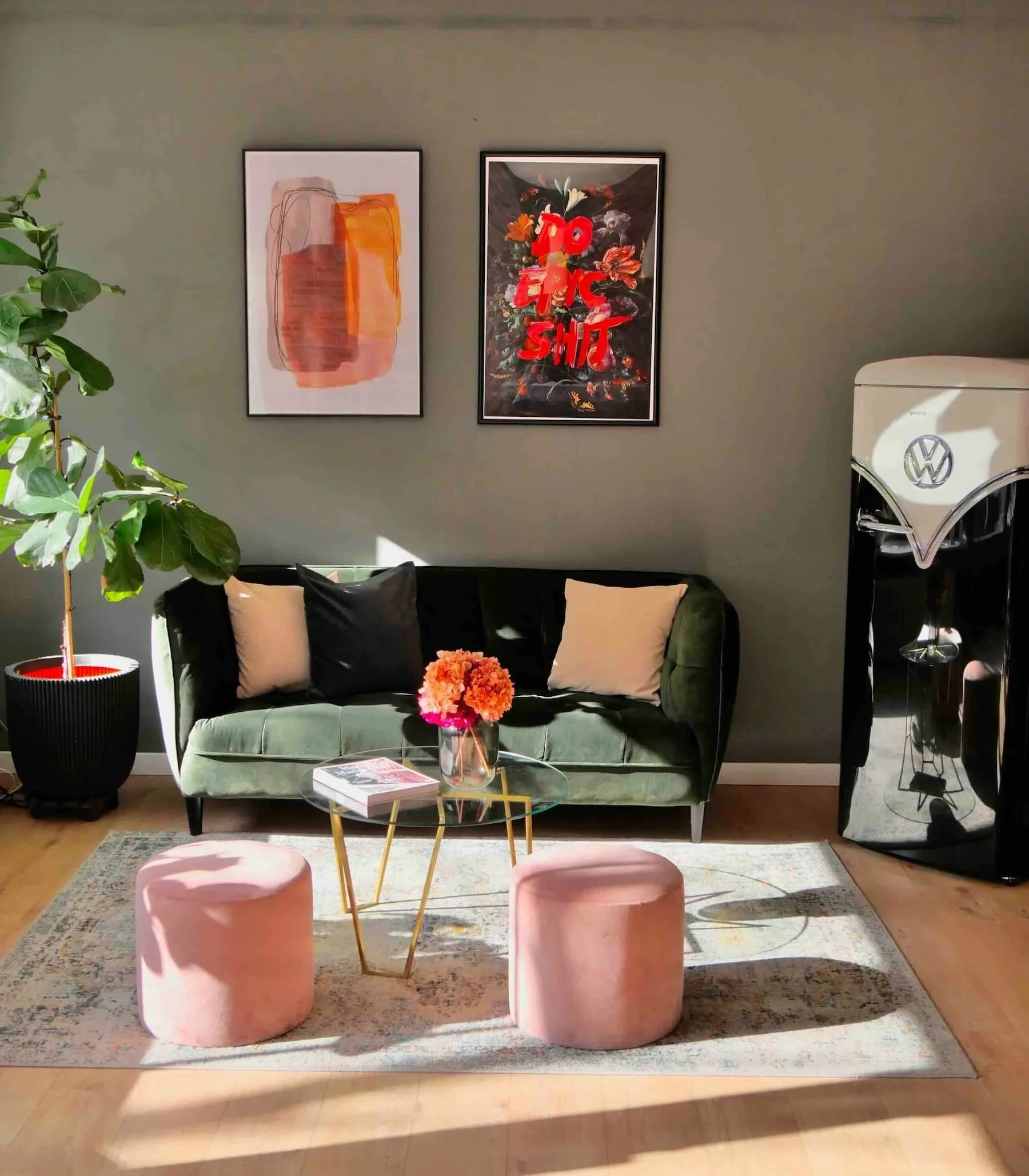 Raum mit grüner Couch, Beistelltisch aus Glas, zwei rosa Poufs. Auf der wand hängen zwei abstrakte Bilder