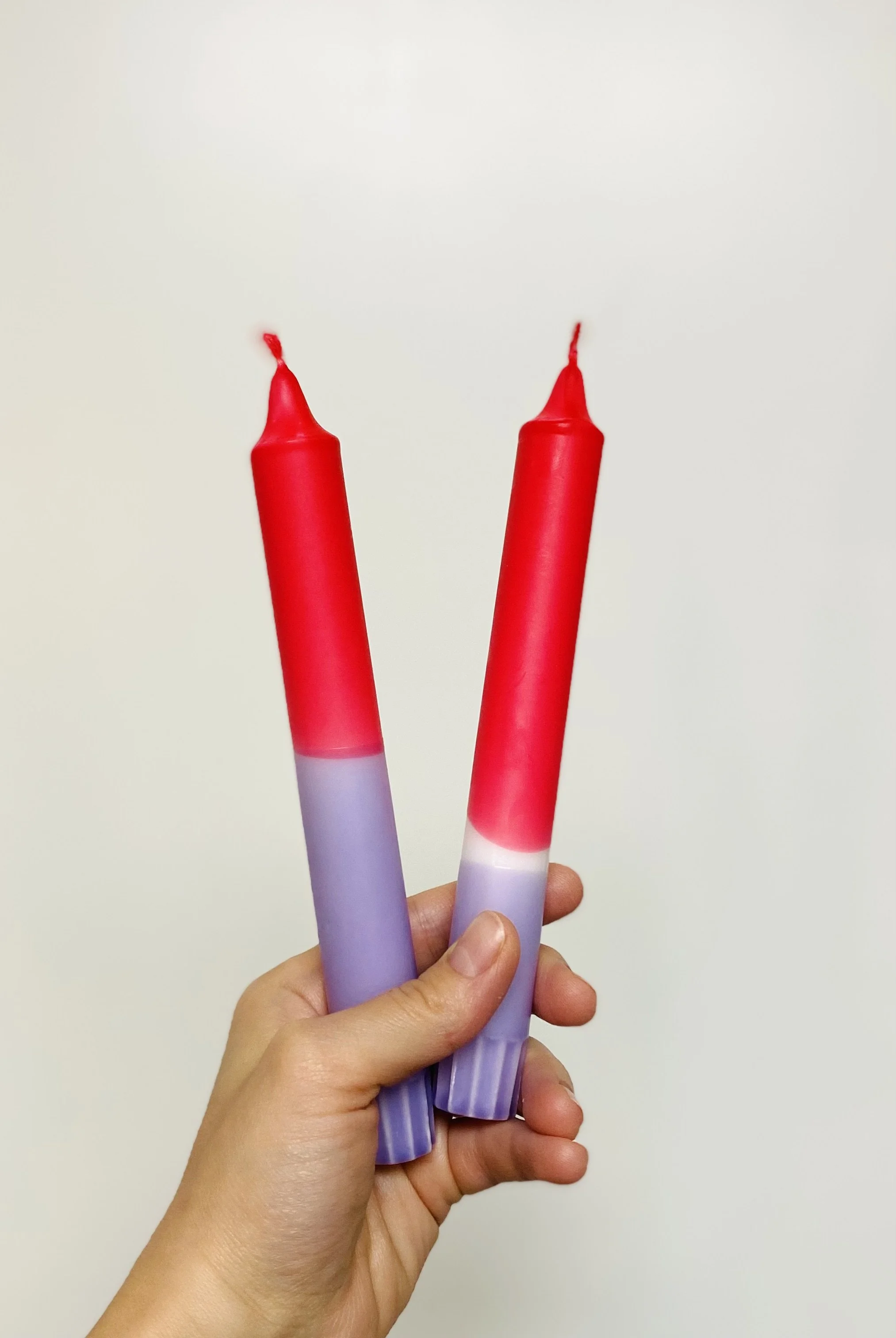 Hand hält zwei rote und lila Kerzen vor einem weißen Hintergrund.
