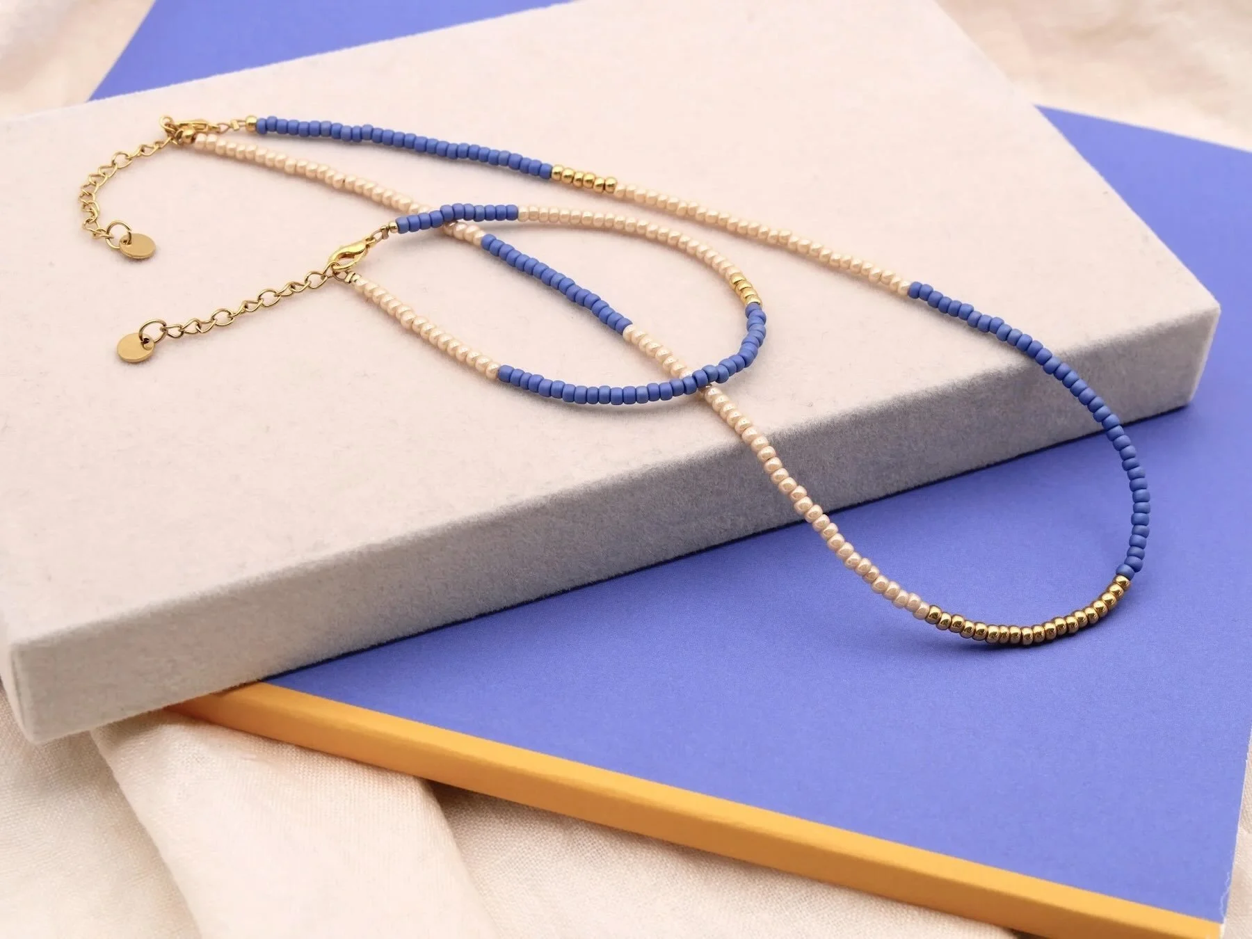 Eine Halskette und ein Armband mit beigen, blauen und goldenen Glasperlen, mit goldenen Karabinern, liegend auf einer beigen Kiste. Hintergrund ist blau.