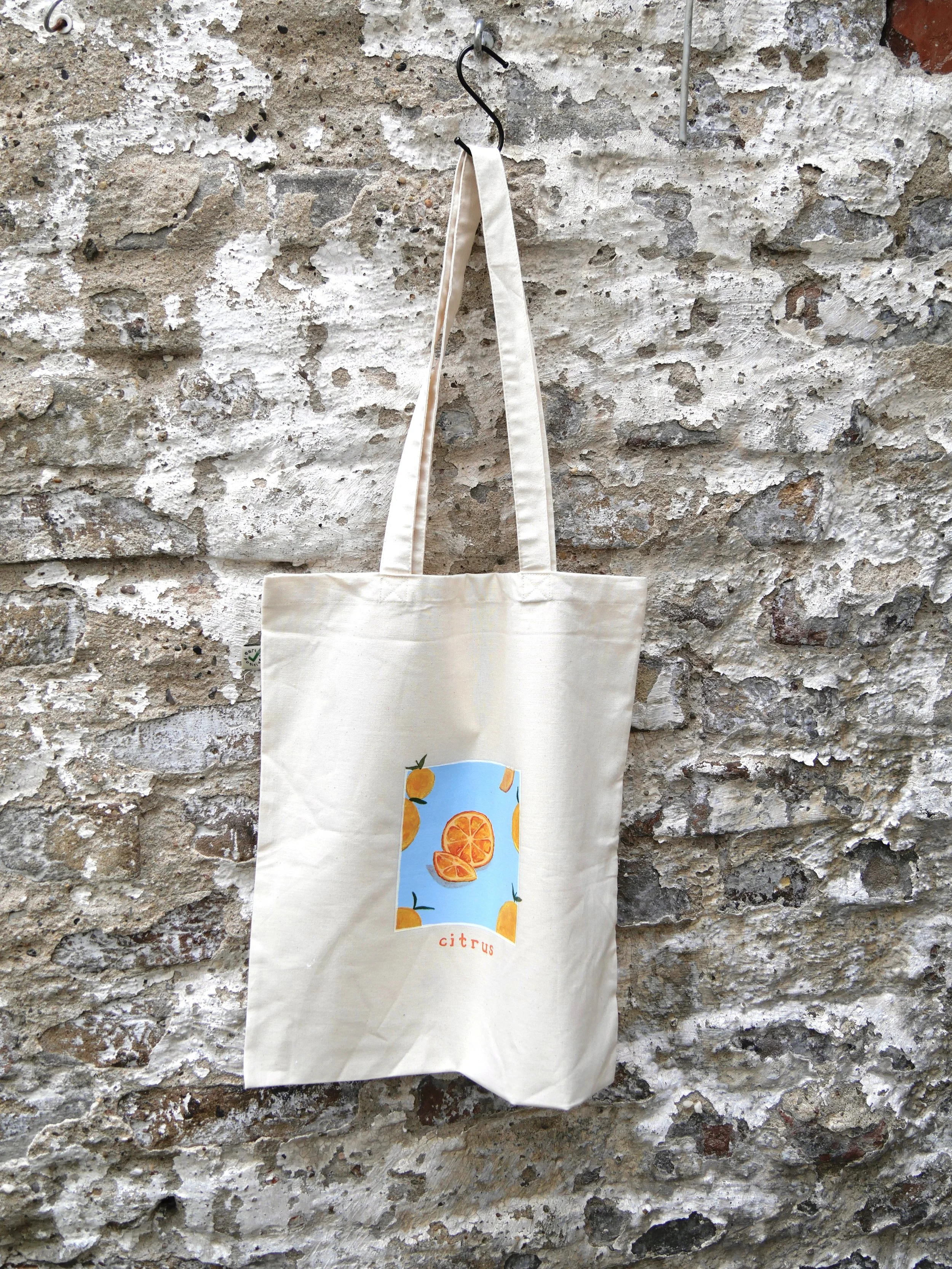 Weiße Leinen Tasche mit gemalten Bild aus Orangenstücken auf hellblauem Vierreck, vor einer Mauer