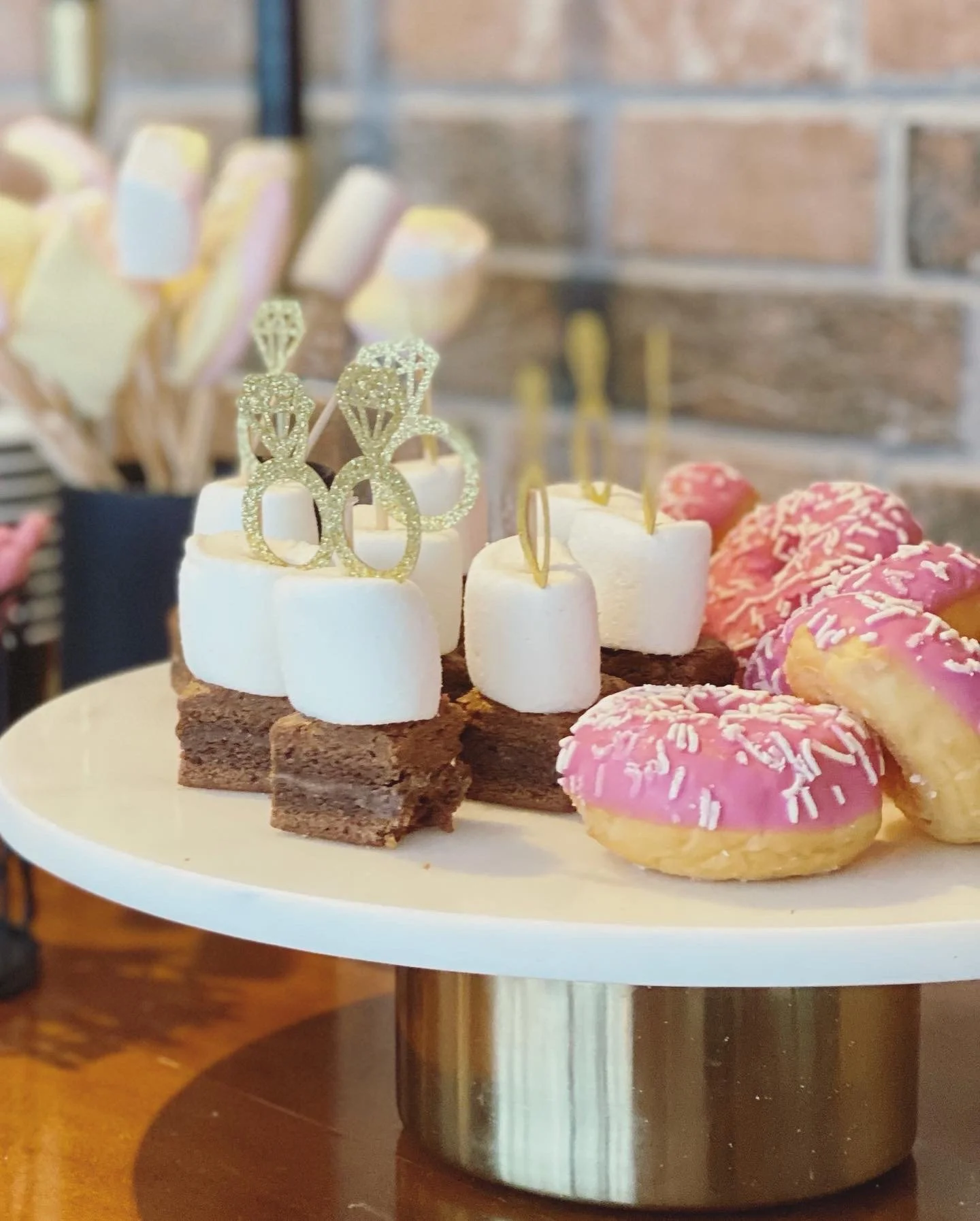 Mehrere kleine Spieße mit Marshmallow und Brownie sowie pinke kleine Doughnuts auf einem Tortenständer mit weißer Platte und breitem goldenem Fuß