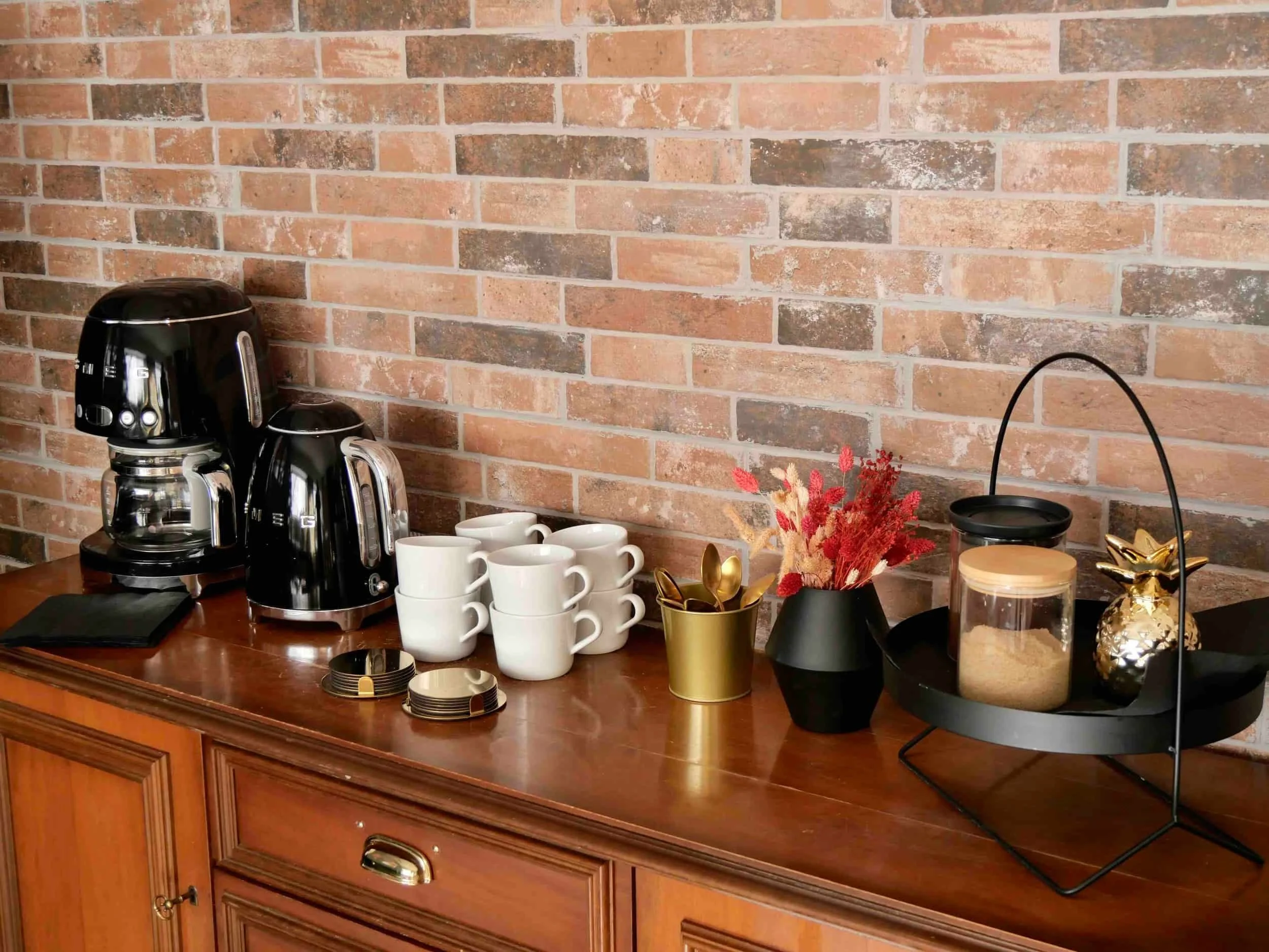 Vintage Kommode mit modernen Kaffemaschine und Wasserkocher in schwarz, Kaffeetassen, Blumenvase, Zucker und weiteren Zubehör, mit Steinen geflieste Wand im Hintergrund