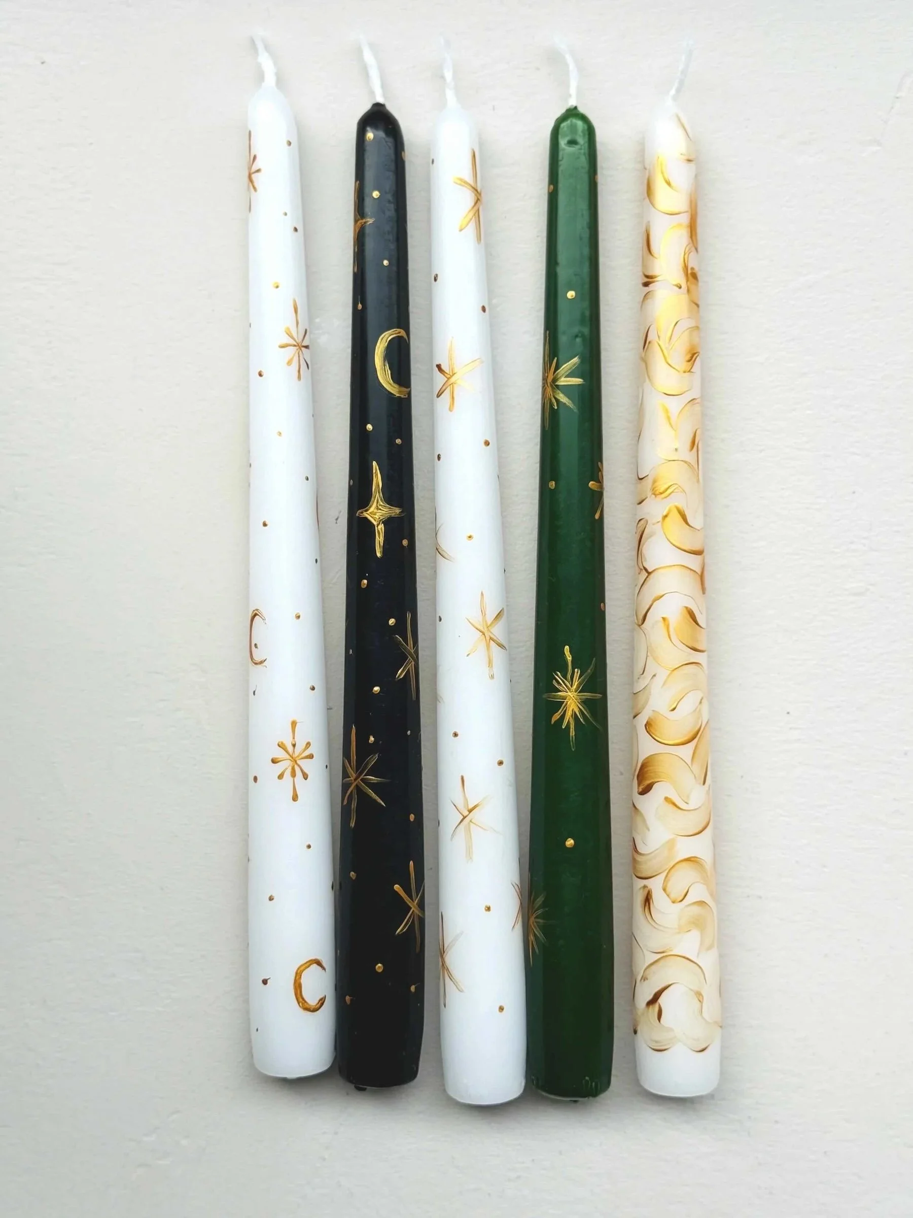 Vier Spitzkerzen in weiß, schwarz, dunkelgrün bemalt mit verschiedenen Weihnachtsmotiven wie Sterne, Mond, Schneeflocken, Wellen in gold
