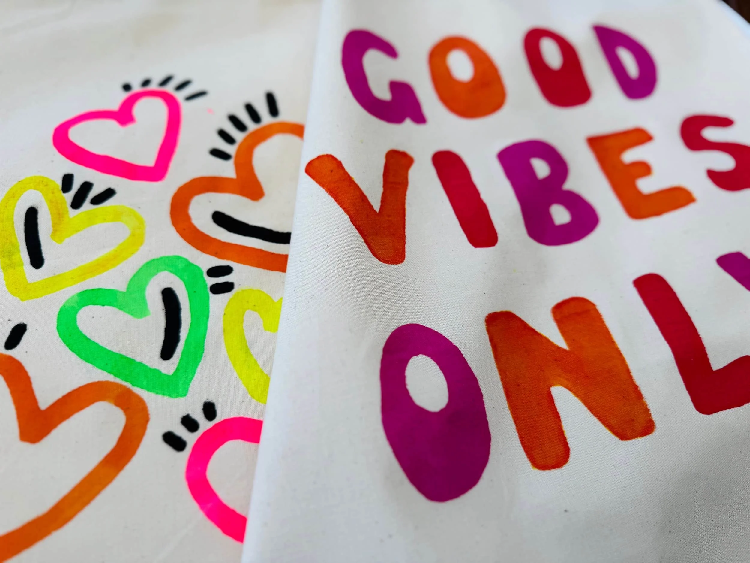 Bunte handgemalte Herzen und die Worte "GOD VIBES ONLY" auf Textiltaschen.