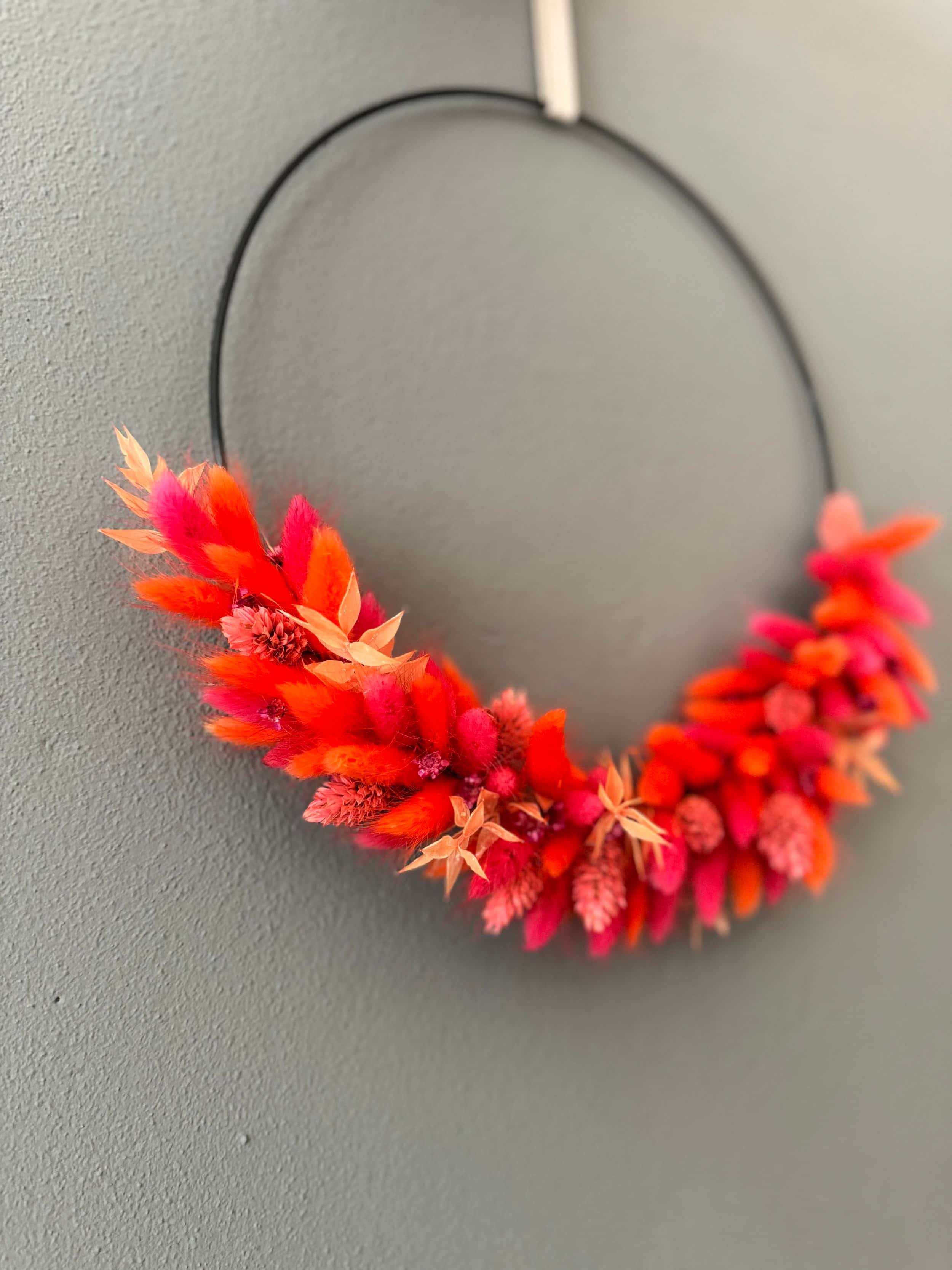 Bunter, halbkreisförmiger Blumenkranz aus roten, orangefarbenen und pinken Trockenblumen hängt an einer grauen Wand.