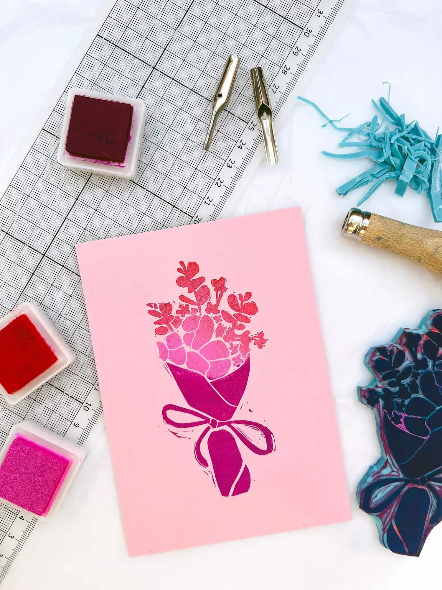 Blockprint eines Blumenstraußes in rosa, rot und pink auf rosanem Papier, umgeben von Stempelkissen, Stempel und Werkzeug
