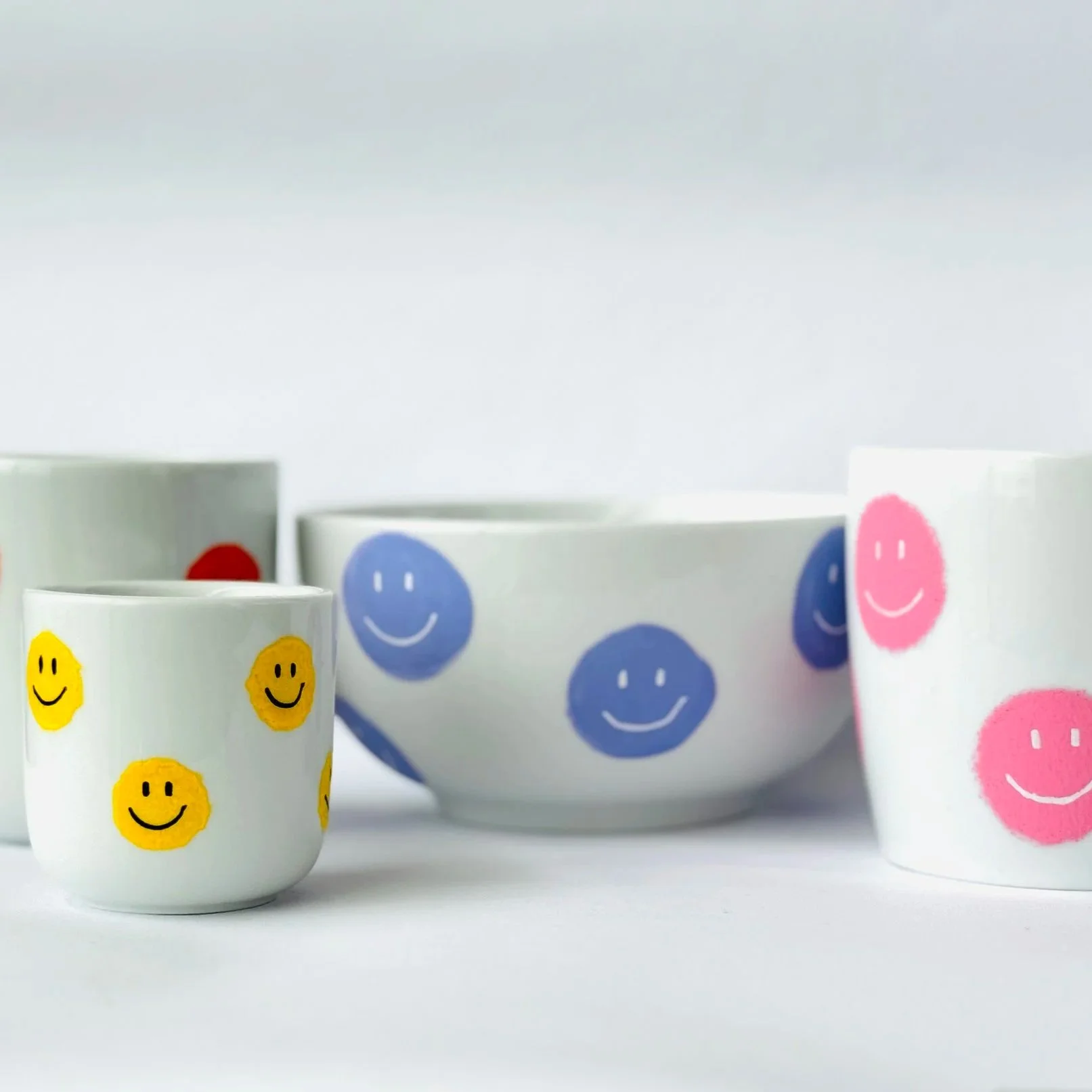 Weiße Tassen und Schalen mit bunten Smileys in Gelb, Rot, Blau und Rosa auf weißem Hintergrund.