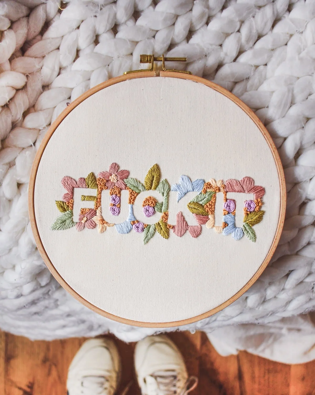 Sticken mit den Wörtern 'FUCK IT' auf einem weißen Stoff, umgeben von bunten, gestickten Blumen und Blättern.