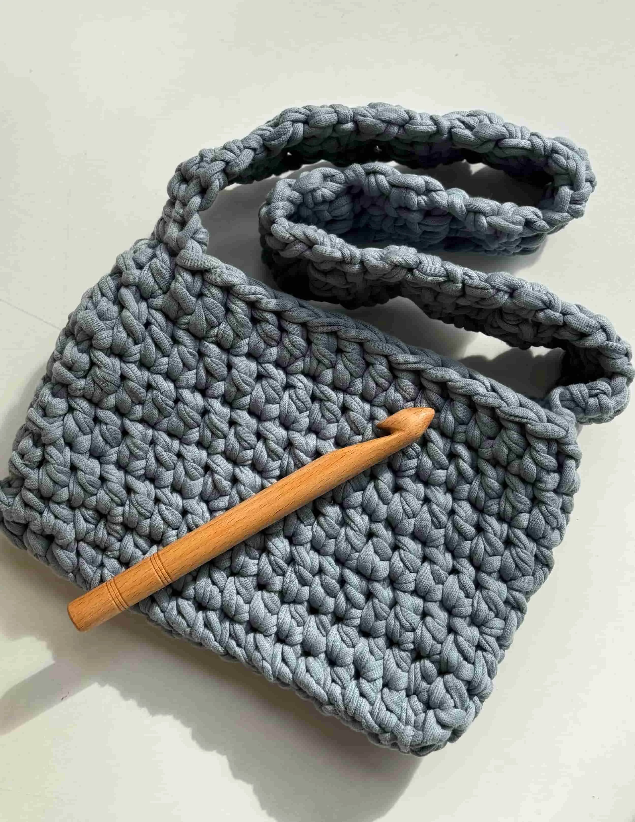 Gehäkelte kleine graue Tasche mit Henkel. Eine Häkelnadel aus Holz liegt auf der Tasche