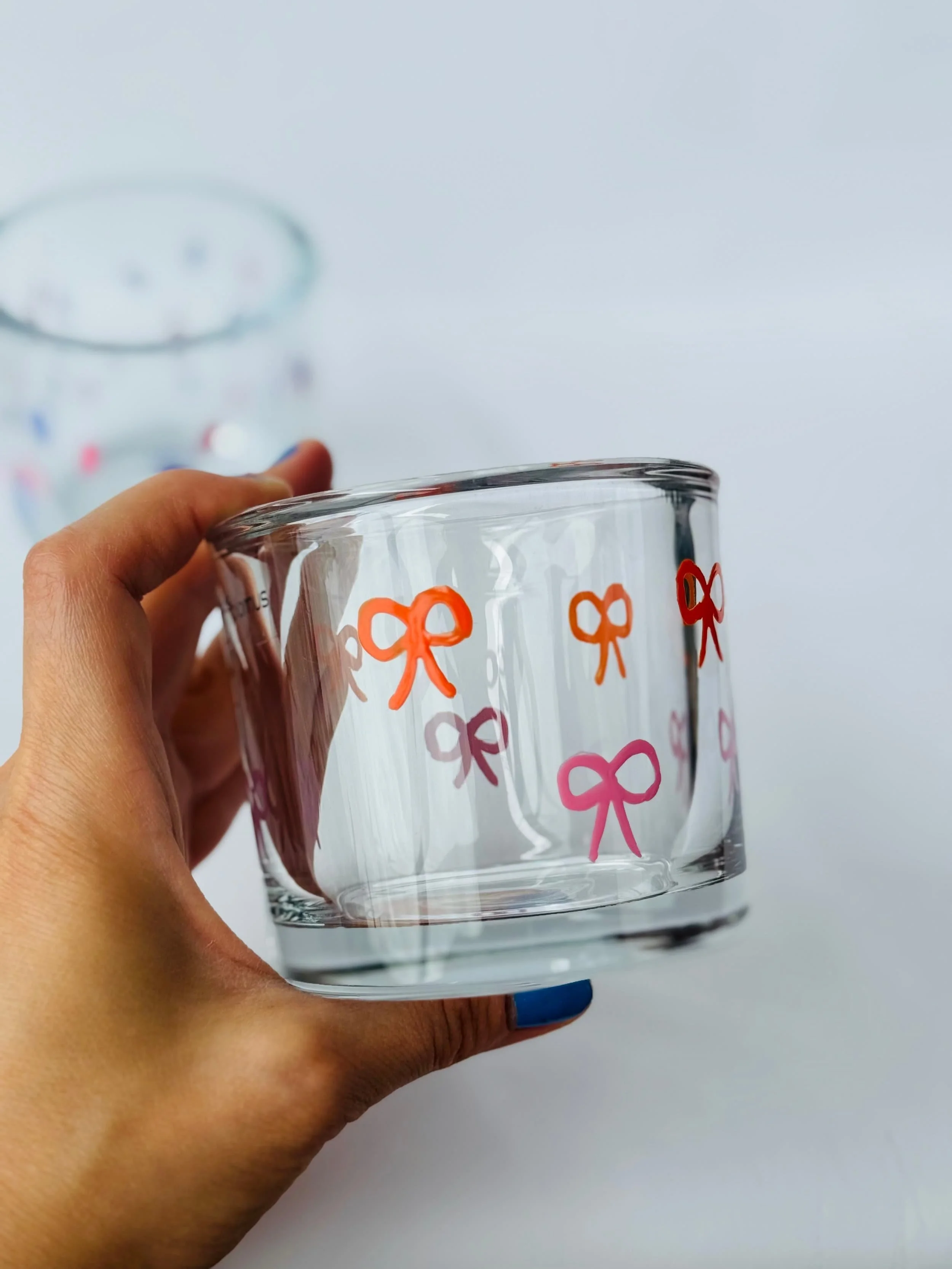 Transparenter Glas mit orangefarbenen und pinkfarbenen Schleifenmuster, gehalten von einer Hand mit blauer Nagelfarbe, vor weißem Hintergrund.