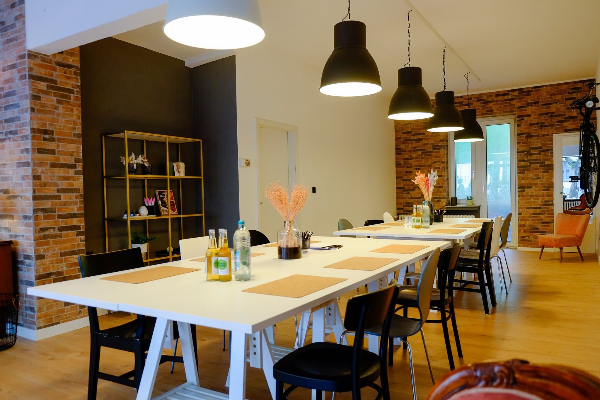 Ein modern eingerichteter Raum mit langen weißen Tischen, schwarzen und weißen Stühlen, floralen Dekorationen, Beleuchtung mit schwarzen Hängelampen, einer Wand mit roten Backsteinmauern, großen Fenstern und einem orangefarbenen Sessel in der Ecke.