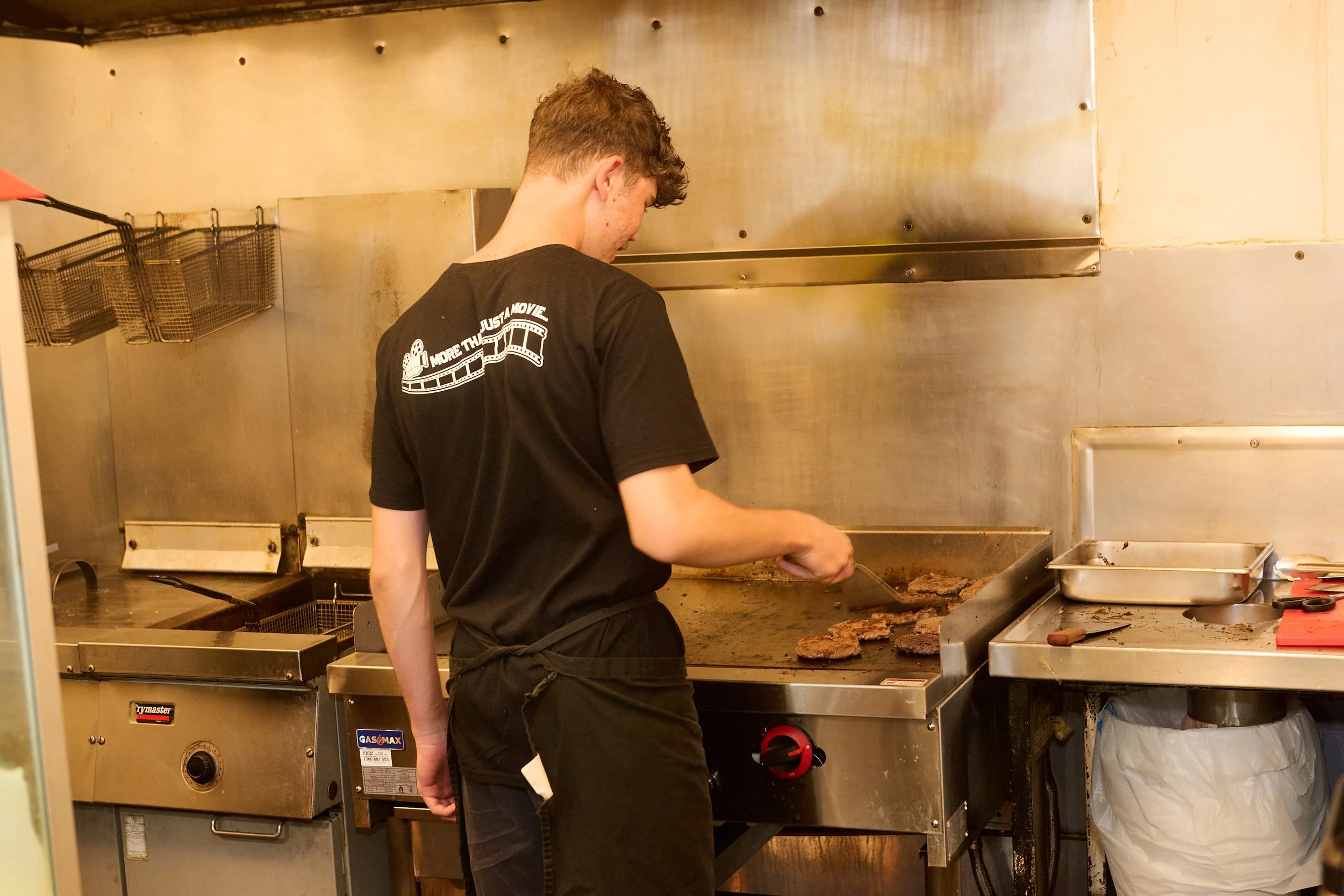 A person cooking meat on a gas grill in a restaurant kitchen. The person is wearing a black shirt with a horizontal logo on the back. The grill is turned on, and the meat is sizzling.