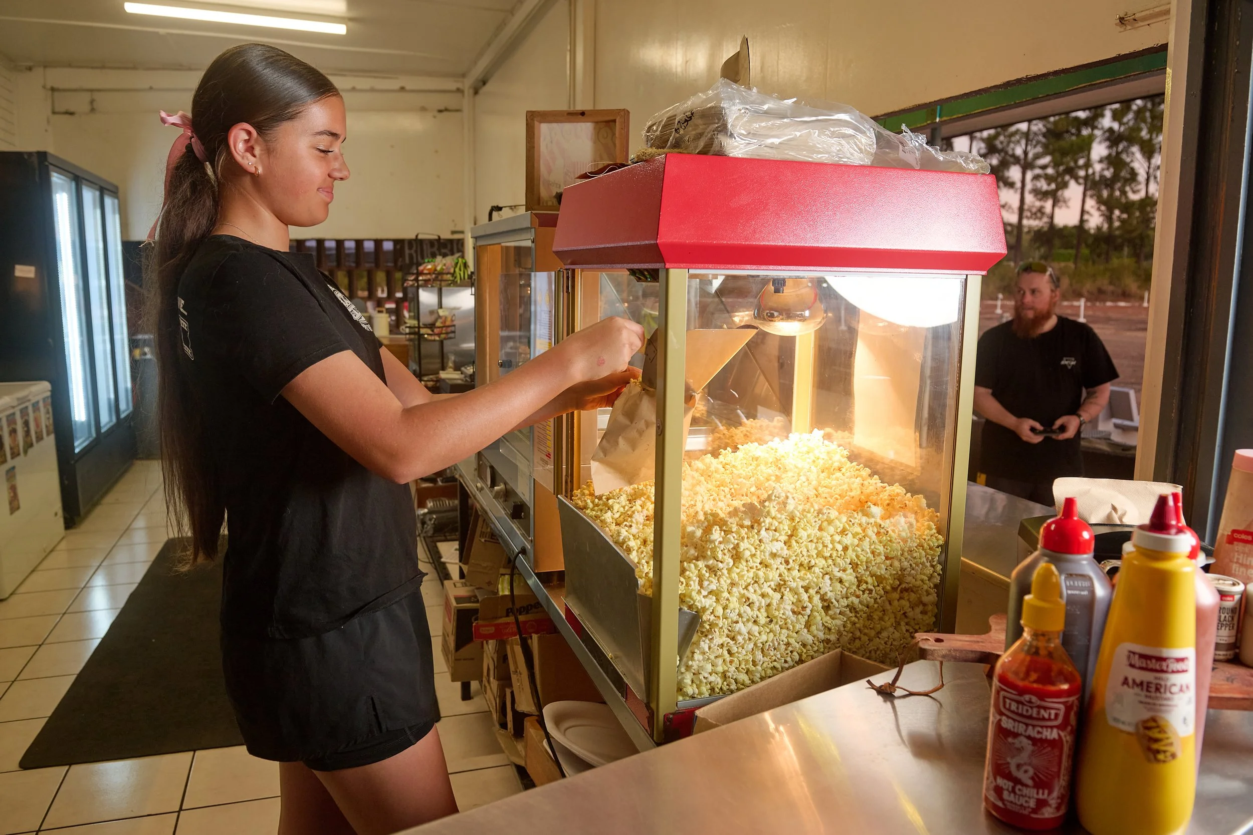 A young woman with long brown hair tied with a pink ribbon, wearing a black t-shirt, is preparing popcorn at a popcorn stand. There is a man with a beard and sunglasses in the background inside the same establishment.
