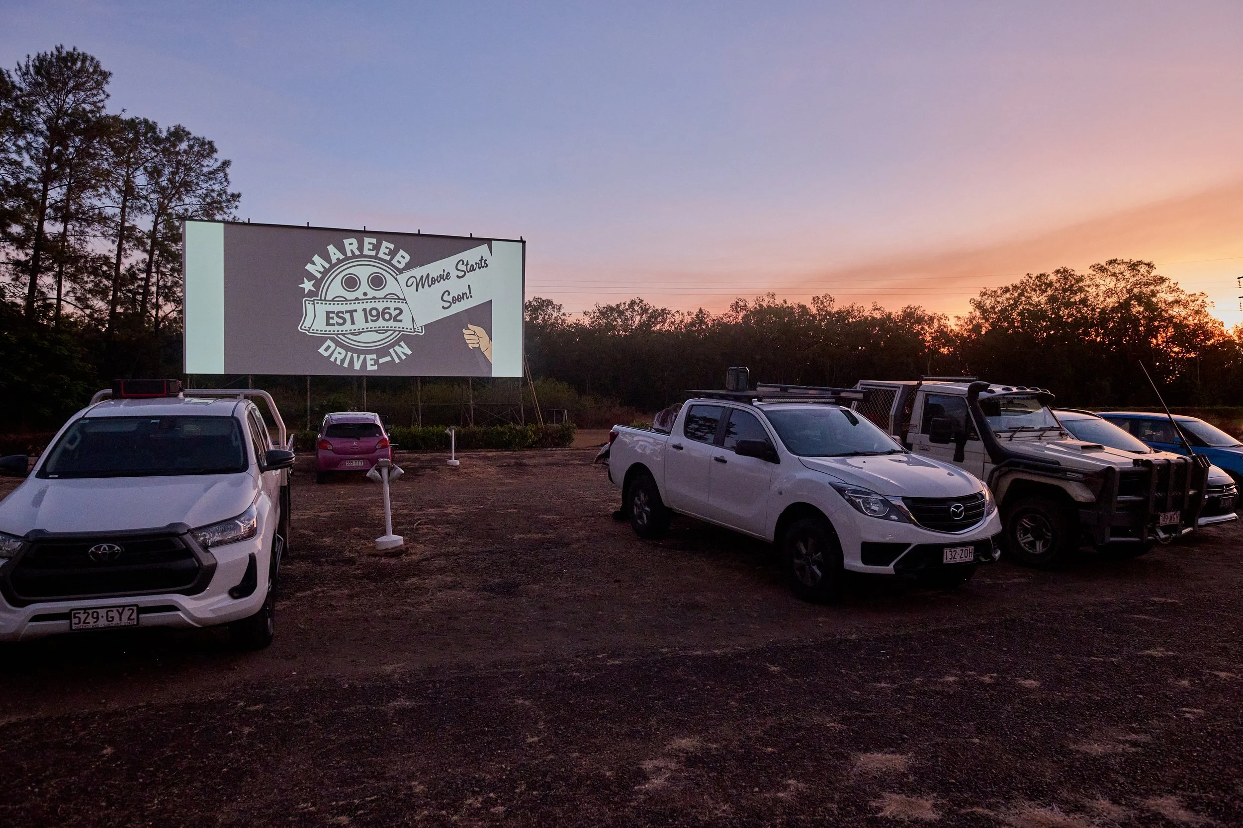 An outdoor drive-in movie theater parking lot at sunset with several parked cars and a large movie screen displaying a vintage-style logo for 'Mareb Drive-In,' established in 1962, announcing that the movie is starting soon.