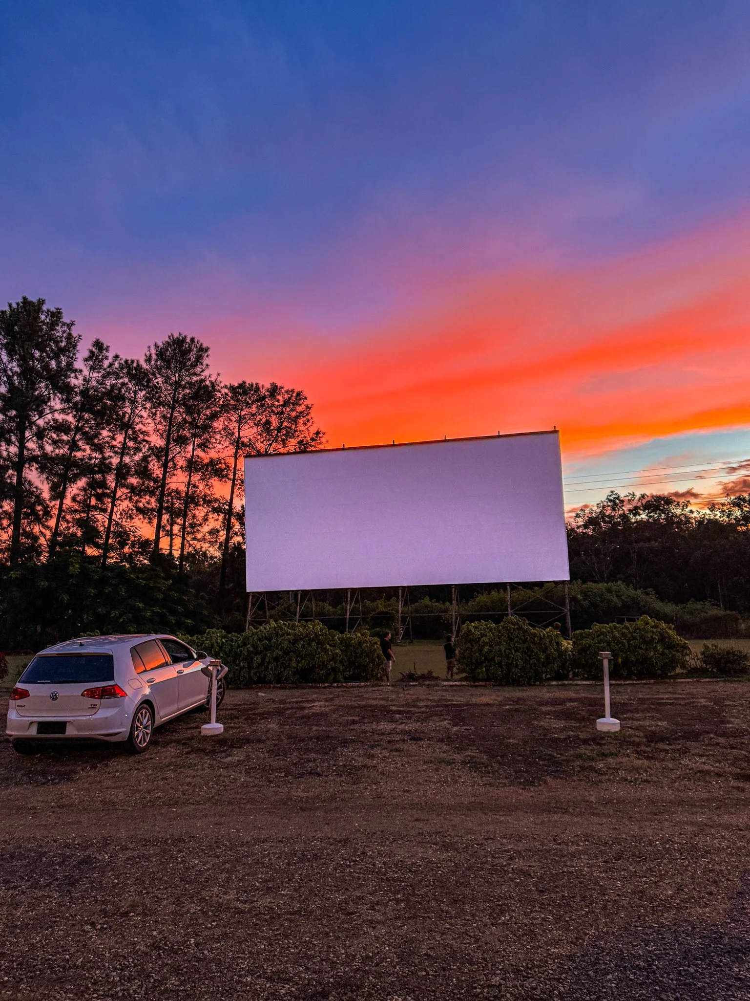 An outdoor movie screen set up at dusk with colorful sunset sky, trees on the left, parked car in front, and a few people near the screen.