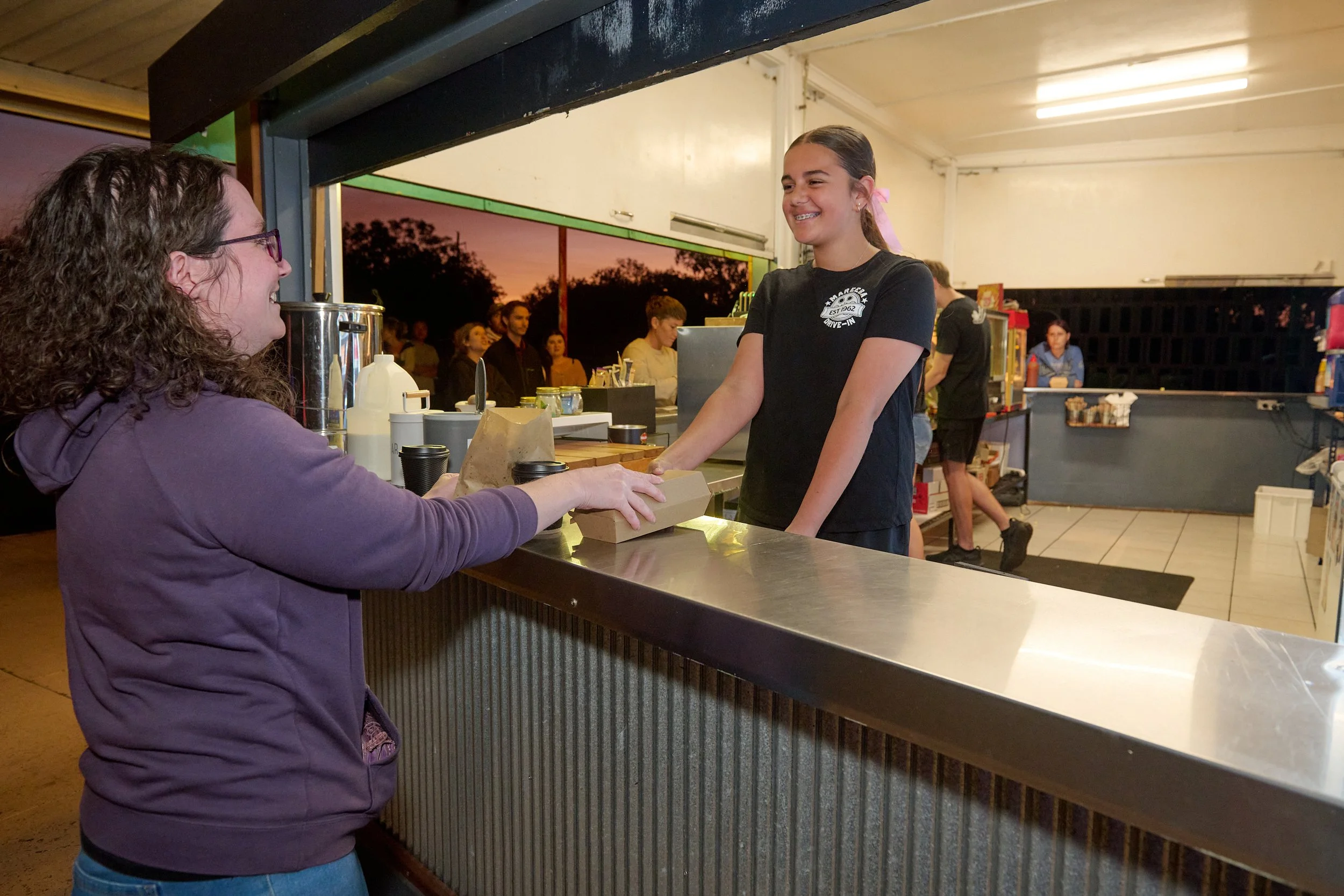 A young girl with braces smiling at a woman at a concession stand at a drive-in theater during sunset.