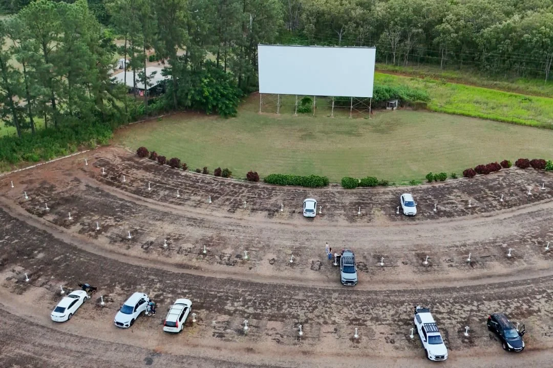An outdoor drive-in movie theater with a large blank screen on a grassy area, surrounded by trees. There are several cars parked in front of the screen on a dirt area, with some people standing near their vehicles. The setting appears to be during daytime.