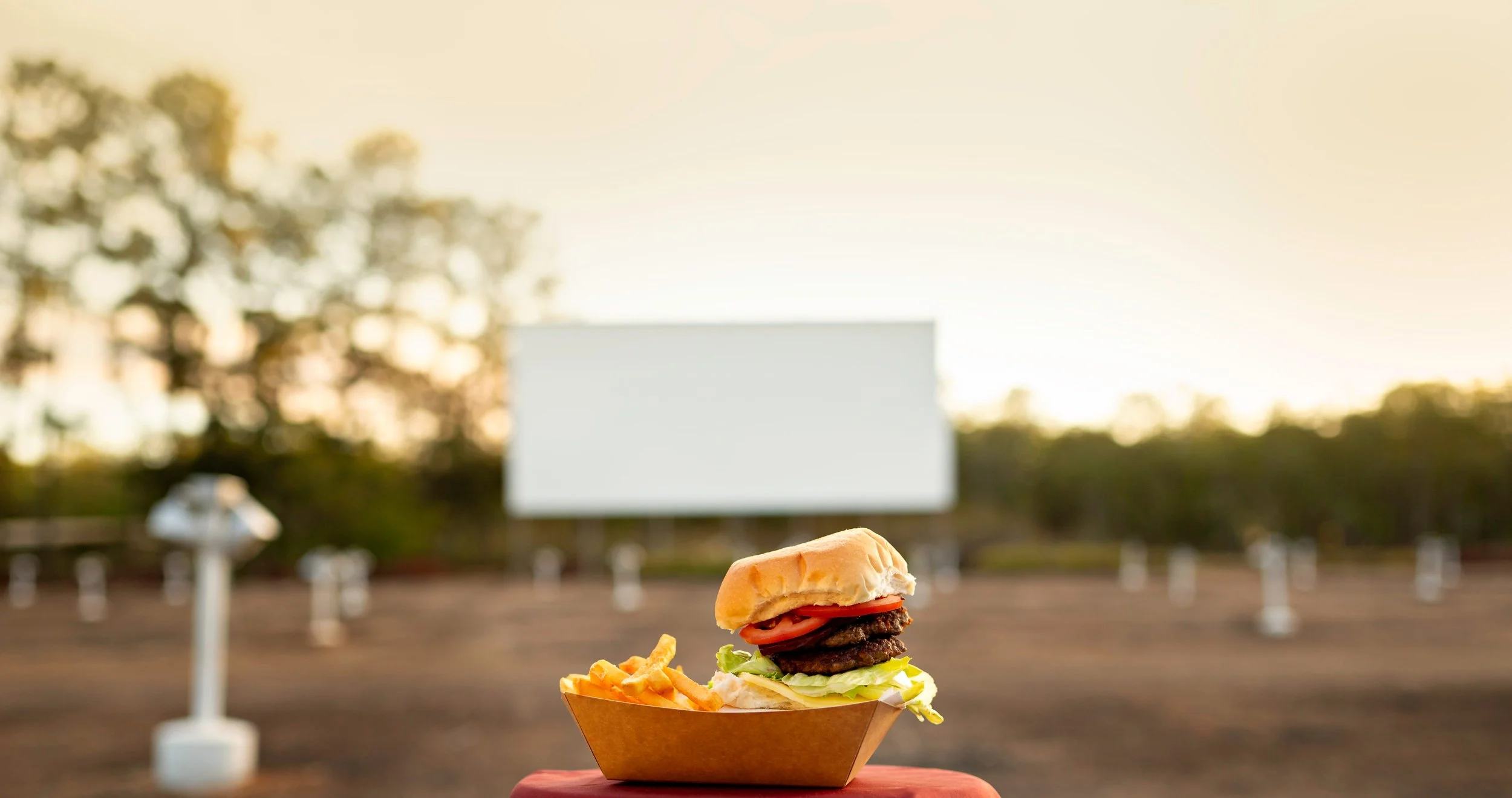 A cheeseburger with lettuce, tomato, and two beef patties served with French fries, outdoors with trees and a drive-in screen in the background.