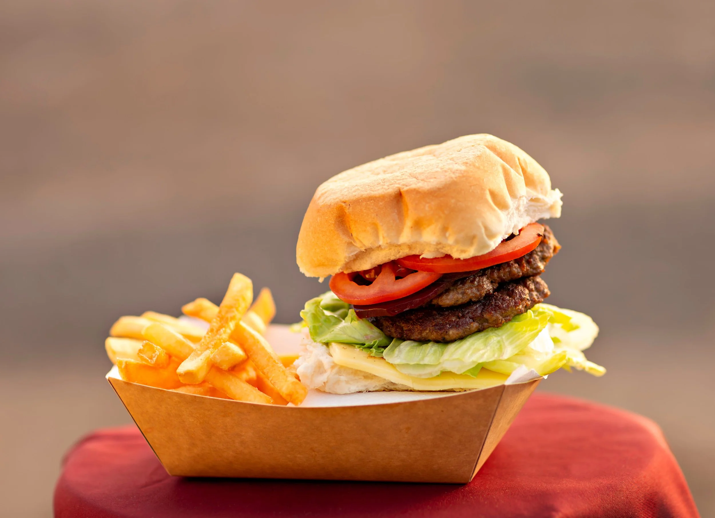 A cheeseburger with lettuce, tomato, pickles, onions, and two beef patties served in a paper food tray, accompanied by French fries on the side.