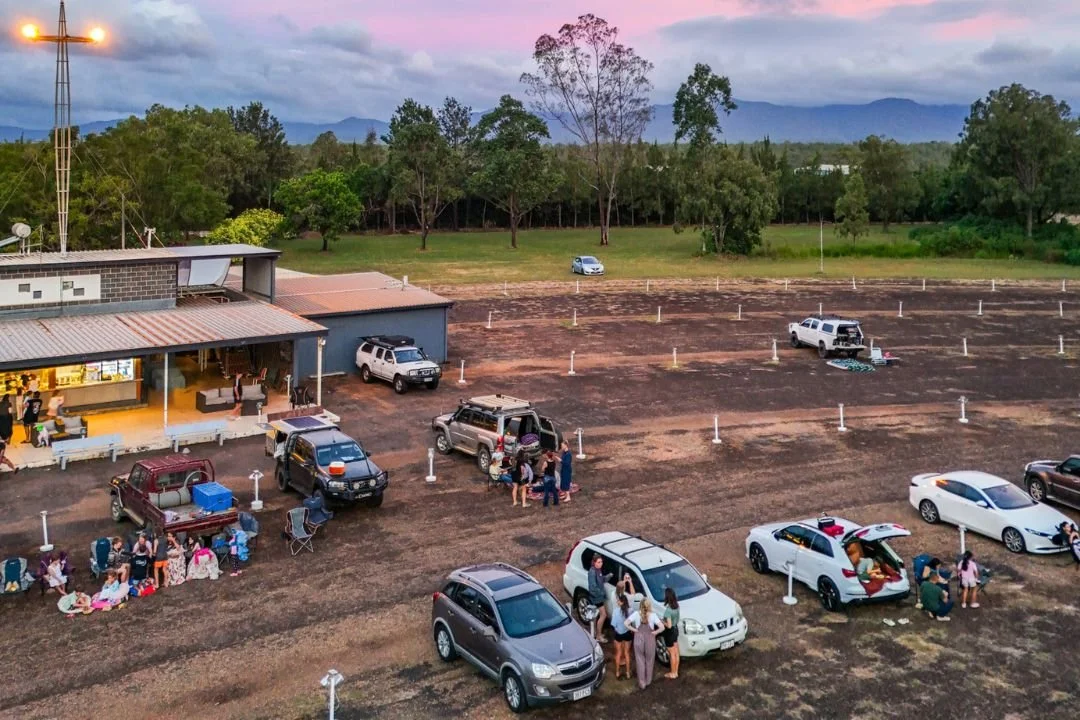 Outdoor gathering with cars parked near a building, people socializing, some sitting on the ground, and others around vehicles outdoors on a dirt lot with trees and mountains in the background during sunset.