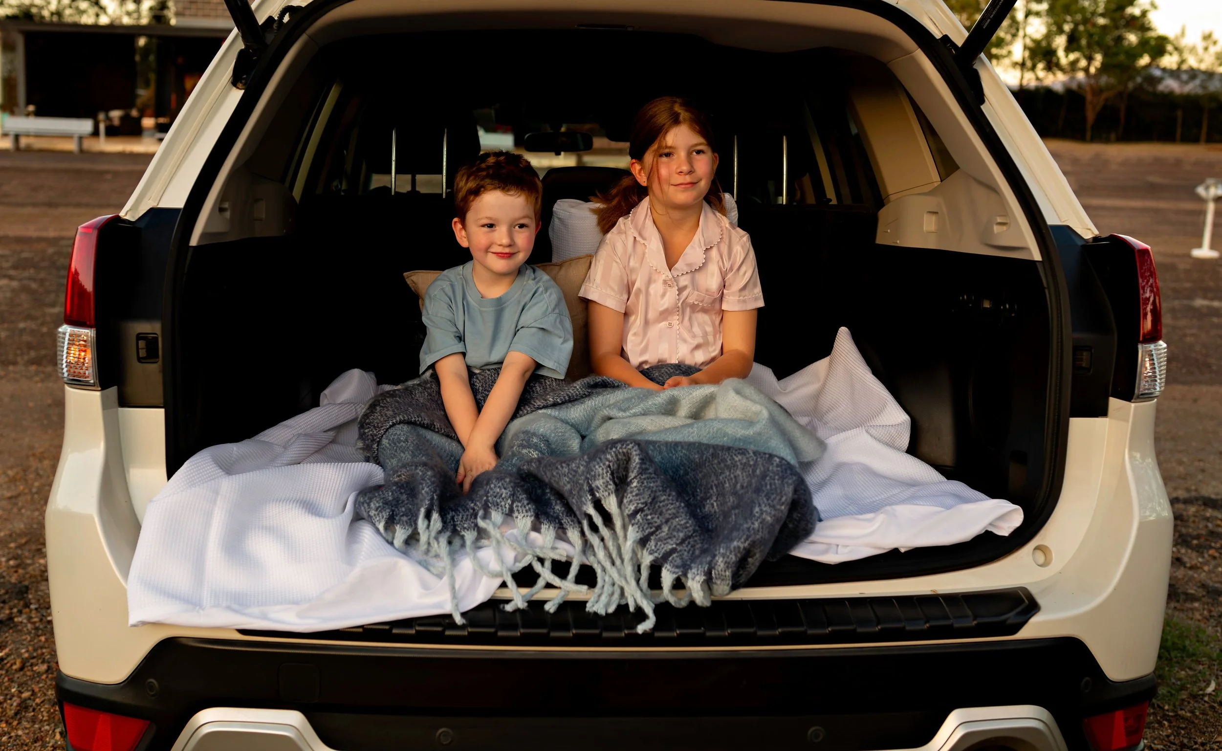 Two children, a boy and a girl, sitting in the back of an open SUV. They are on a blanket with blankets on their laps, outdoors during sunset or early evening.