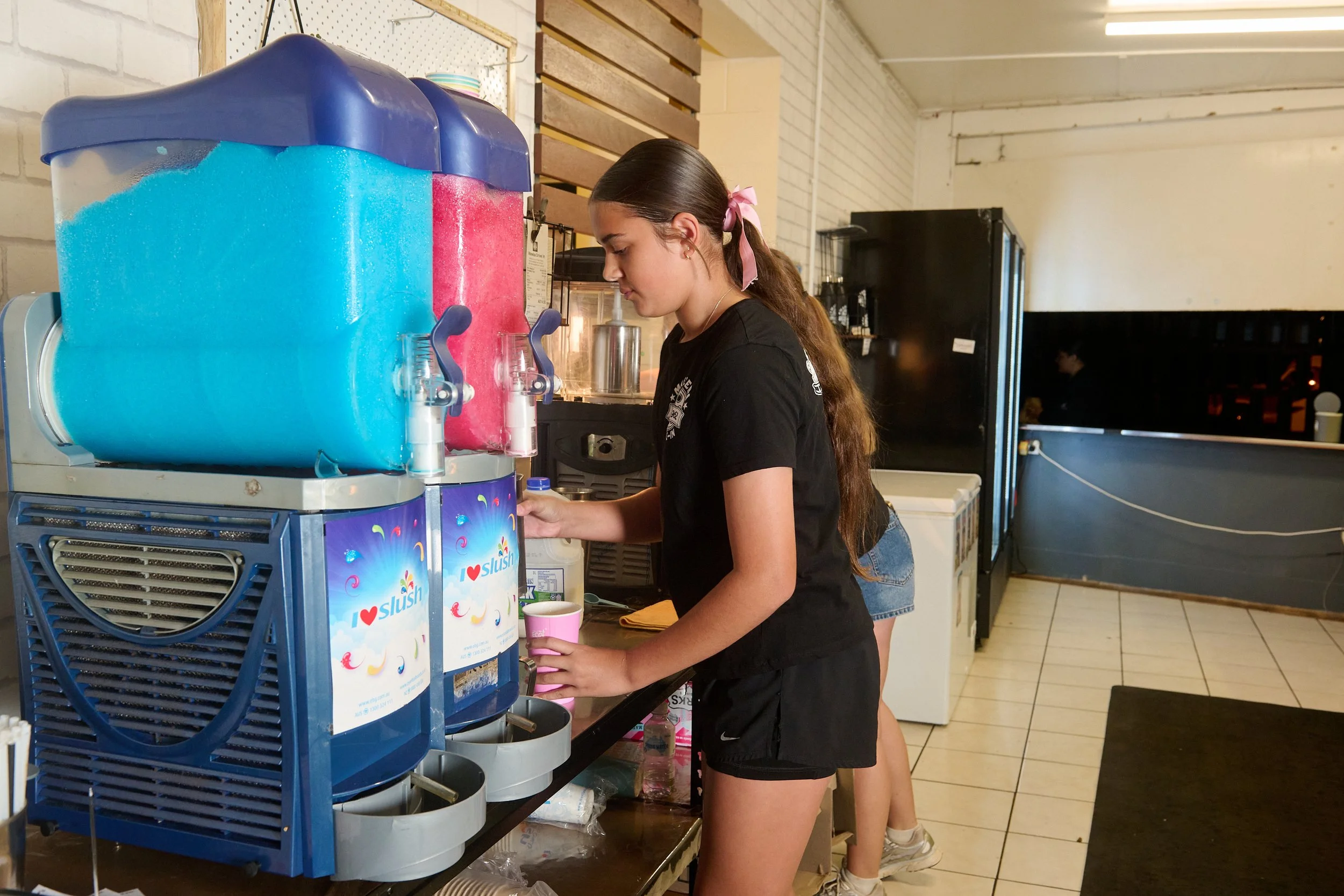 A young woman with long brown hair tied with a pink ribbon, wearing a black T-shirt and black shorts, pouring pink slush from a soda fountain machine into a pink cup at a cafe or ice cream shop.