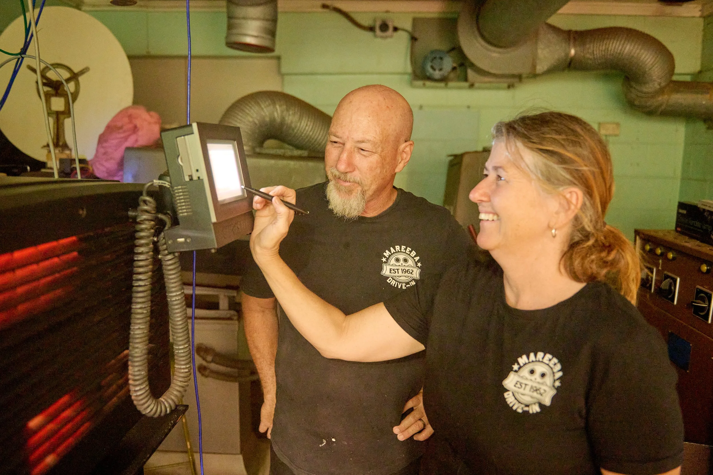 A man and a woman are in a workshop or garage, looking at a small monitor. The man has a bald head with a beard, and the woman has red hair tied back. Both are smiling and wearing black T-shirts with a logo that says 'MAREEBAN DRIVE-IN EST 1962.' The