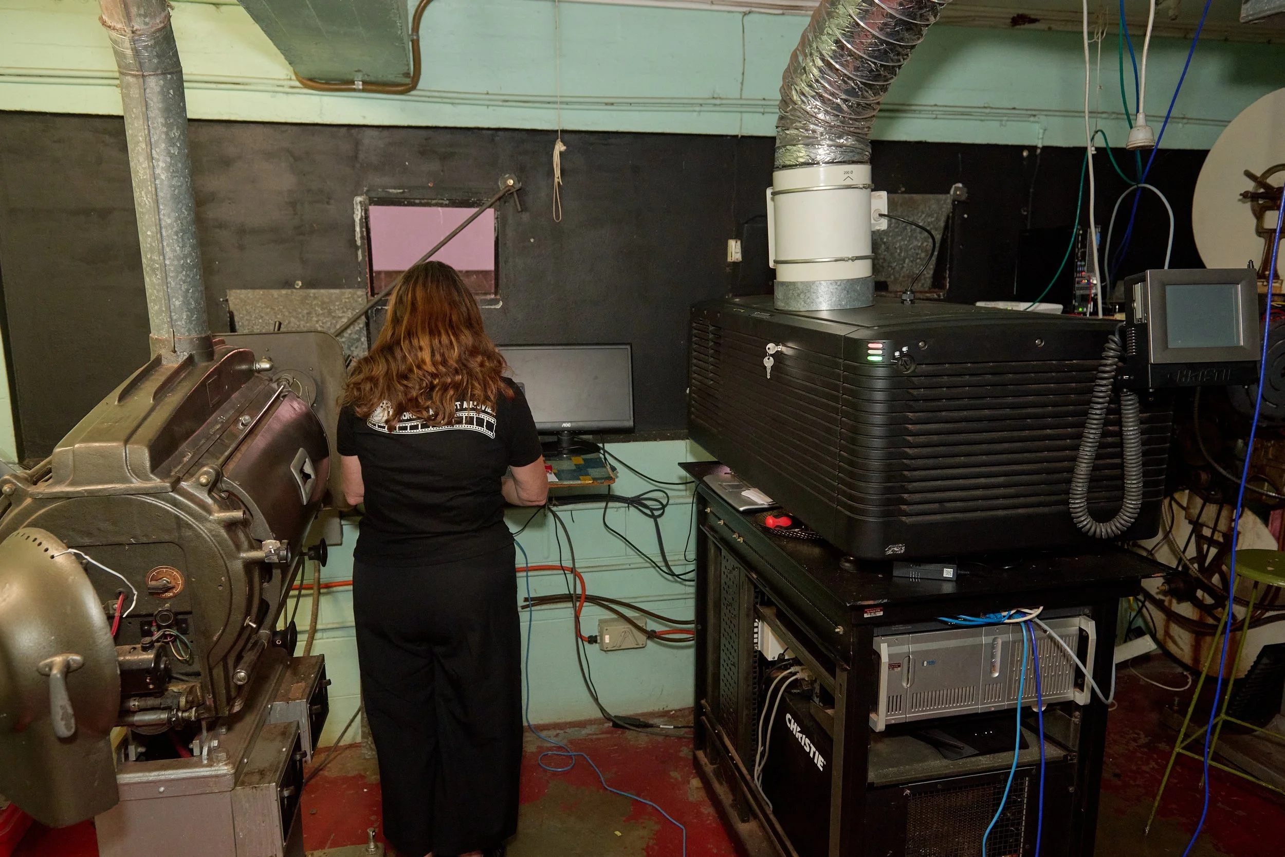 A person with long, wavy brown hair is working on electronic or technical equipment in a workshop or laboratory. The setup includes large machinery, wires, and a computer monitor, in a room with a black and green wall and a red floor.