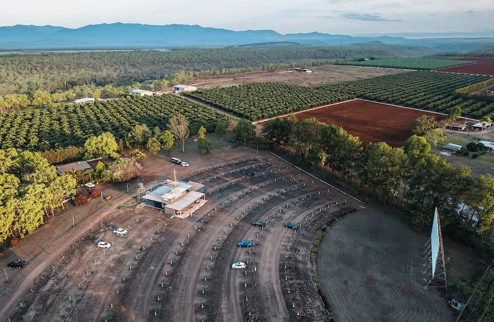 A large outdoor movie theater with a curved rows of seats, surrounded by trees, with a building at the front and a large blank screen, in a rural area with farmland and mountains in the background.