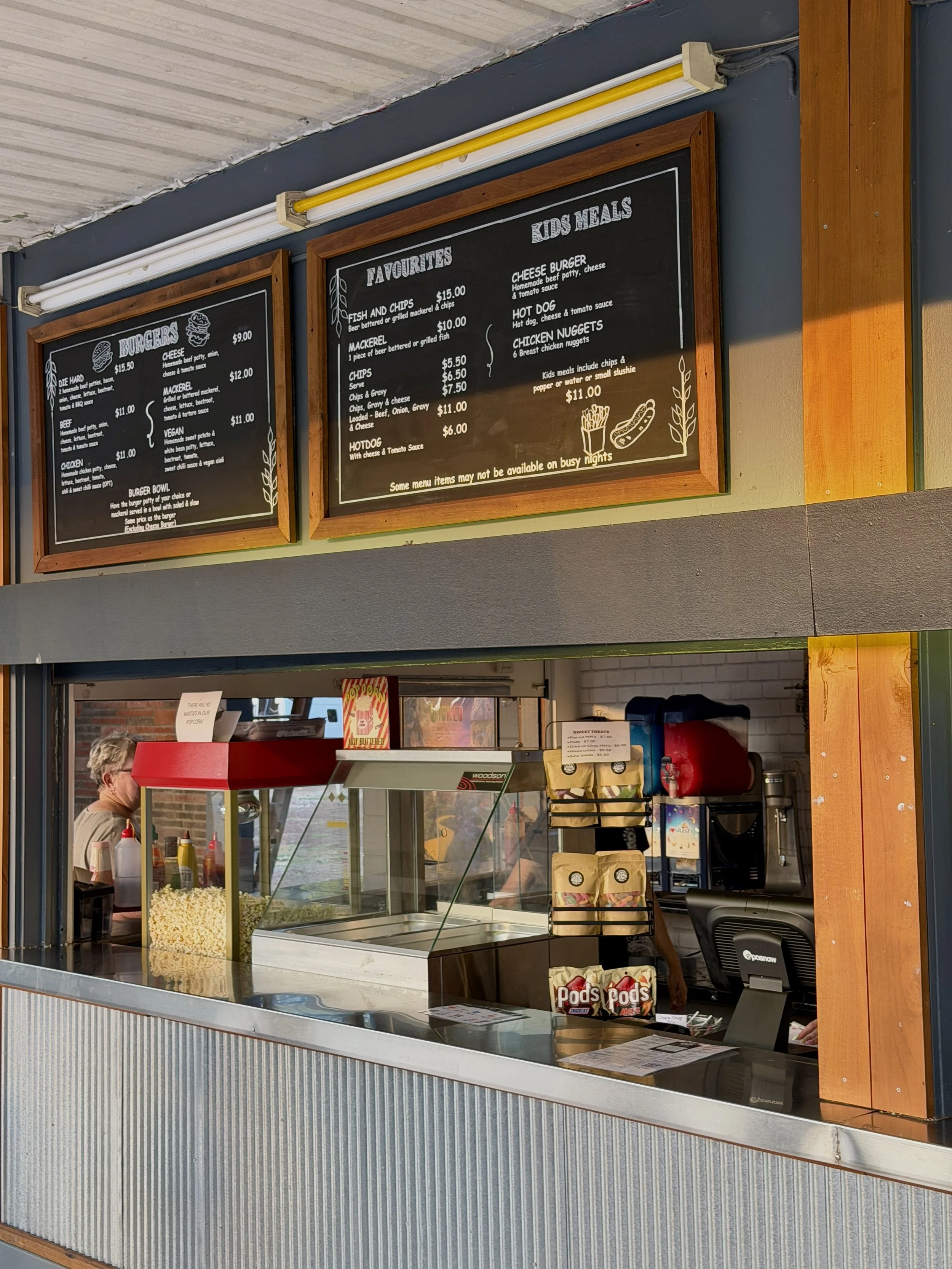 Food stand with menu boards displaying various burger options and kids meals, counter with popcorn and snacks, and a worker preparing food in the background.