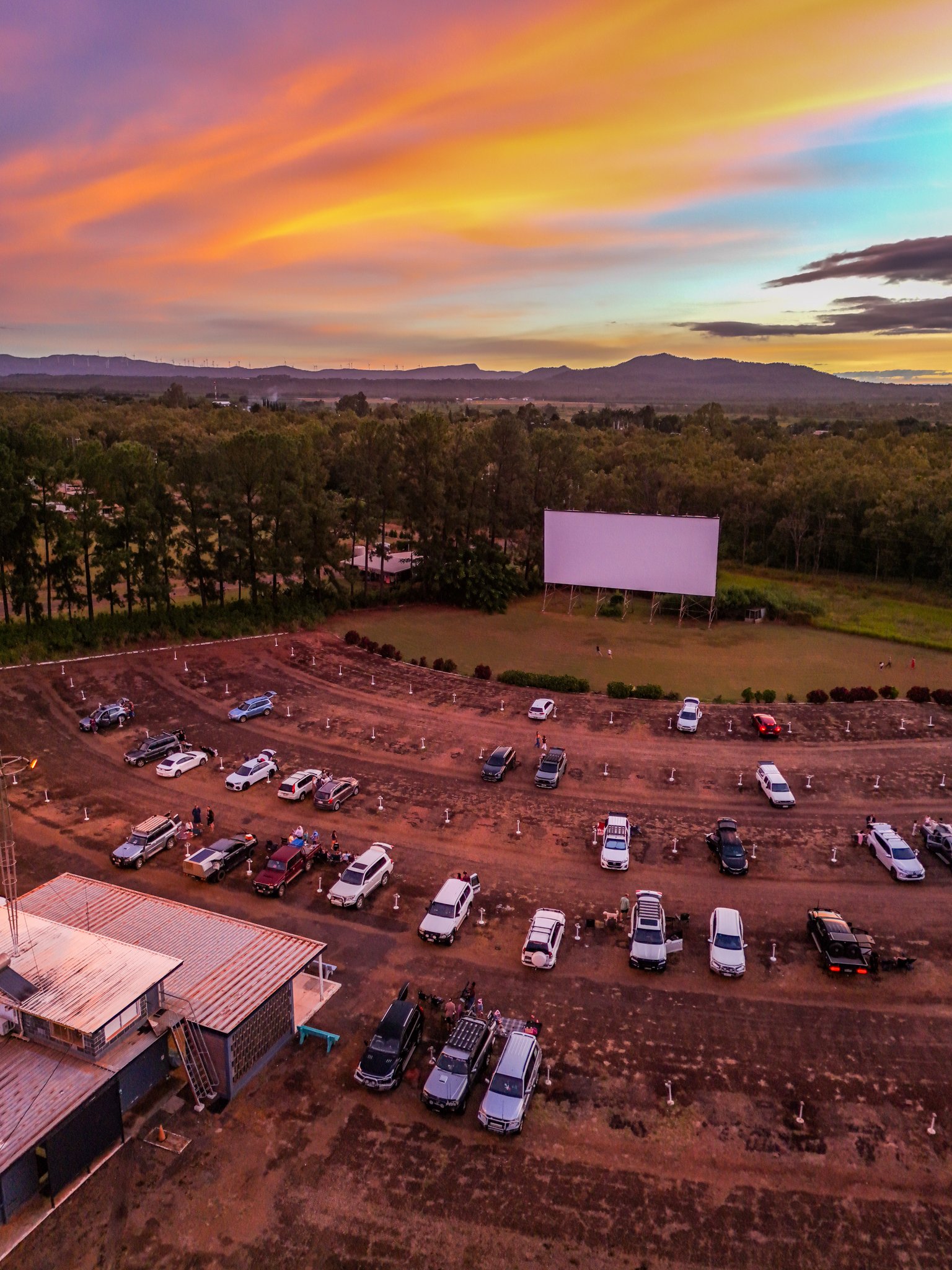 A parking lot filled with cars, next to a small building with a red roof, and a large open field with a big blank white outdoor movie screen, during sunset with colorful clouds in the sky. Trees and hills are in the background.