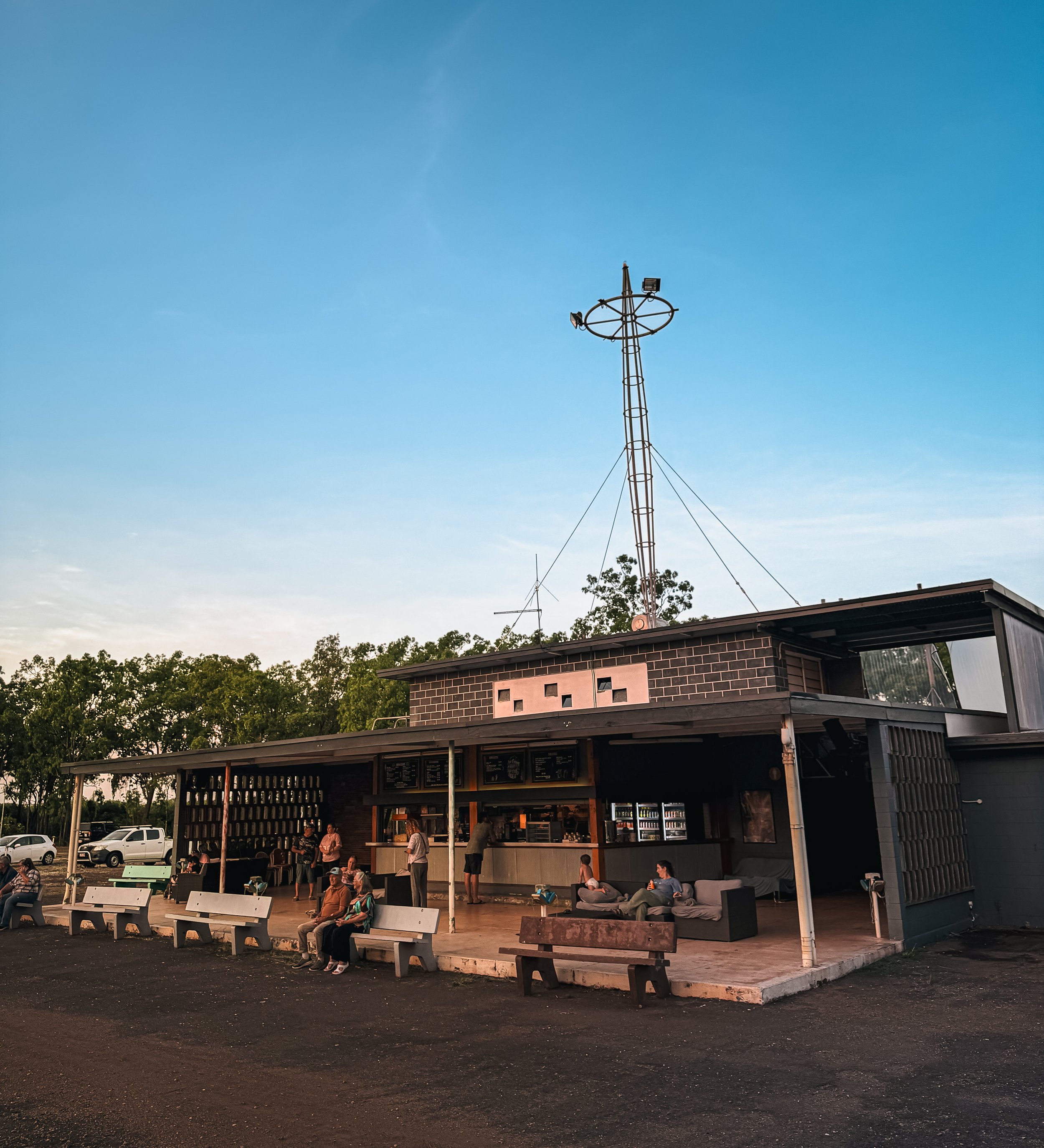 An outdoor caféwith a modern brick building and a large antenna structure on top, under a blue sky with some clouds. There are people sitting on benches and chairs outside, some waiting, some chatting, and some looking at their phones. Trees and park