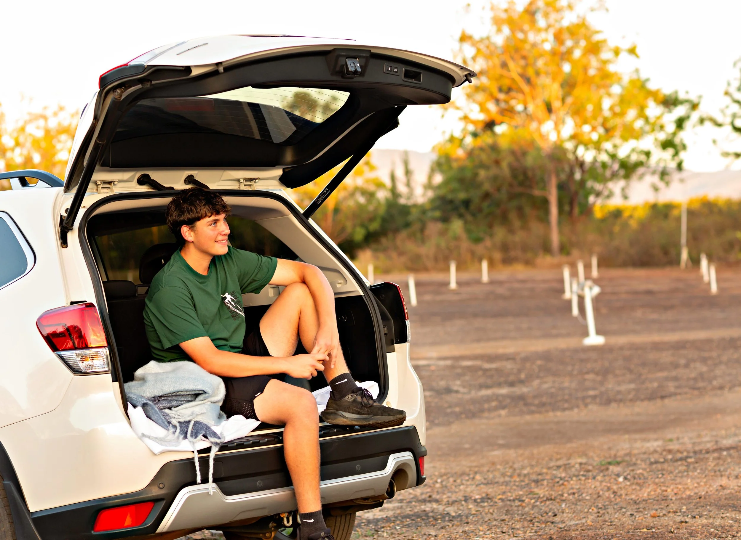 A young man with curly hair sits in the open trunk of a white SUV, smiling and looking to the side, with autumn trees in the background.