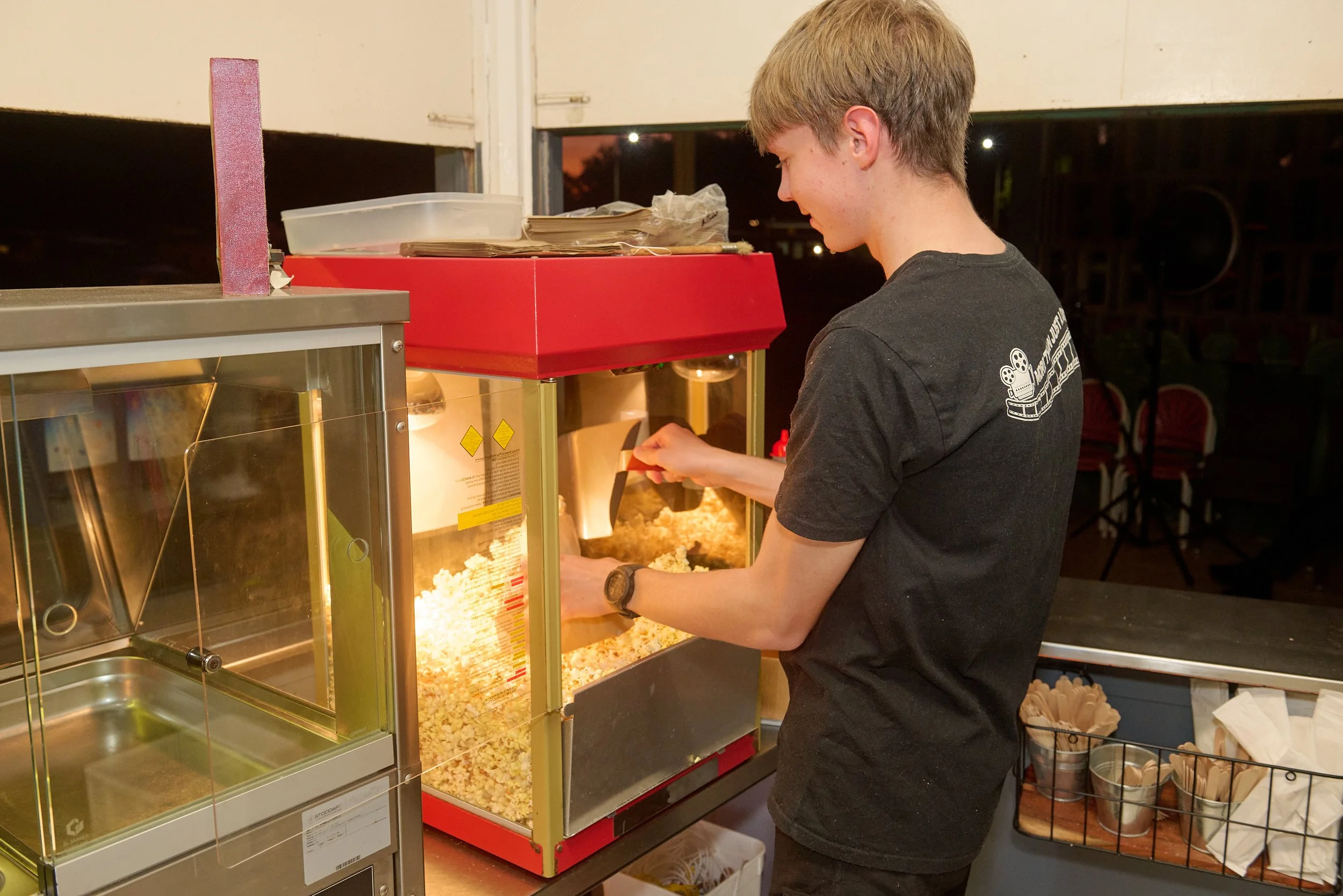 A young man with light brown hair wearing a black t-shirt serves popcorn at a popcorn machine in a concession stand.