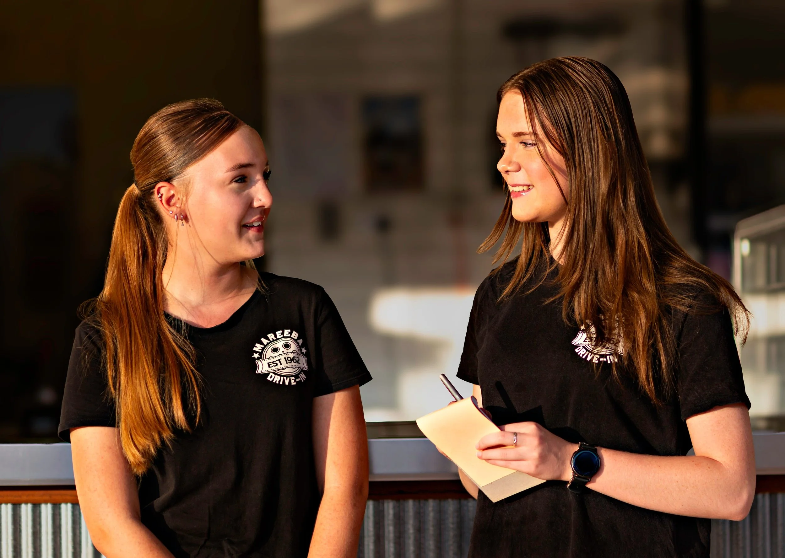 Two young women wearing black T-shirts with a logo engaged in conversation in an indoor setting.