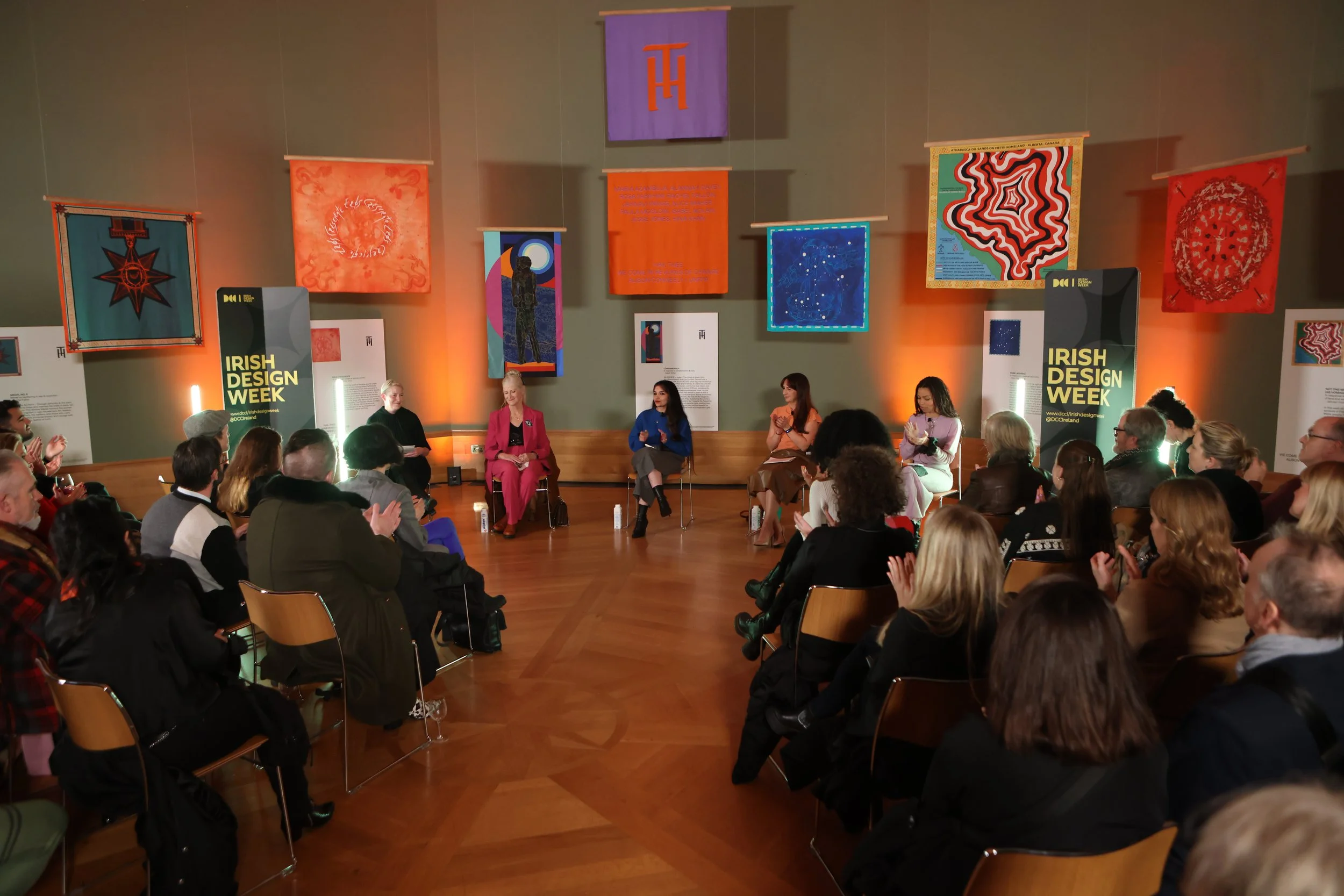 A panel of five women seated on stage at Irish Design Week, with an audience in front. The stage backdrop features colorful banners and artworks, including geometric patterns and celestial motifs. The event appears to be a discussion or presentation in a well-lit art gallery or conference room.