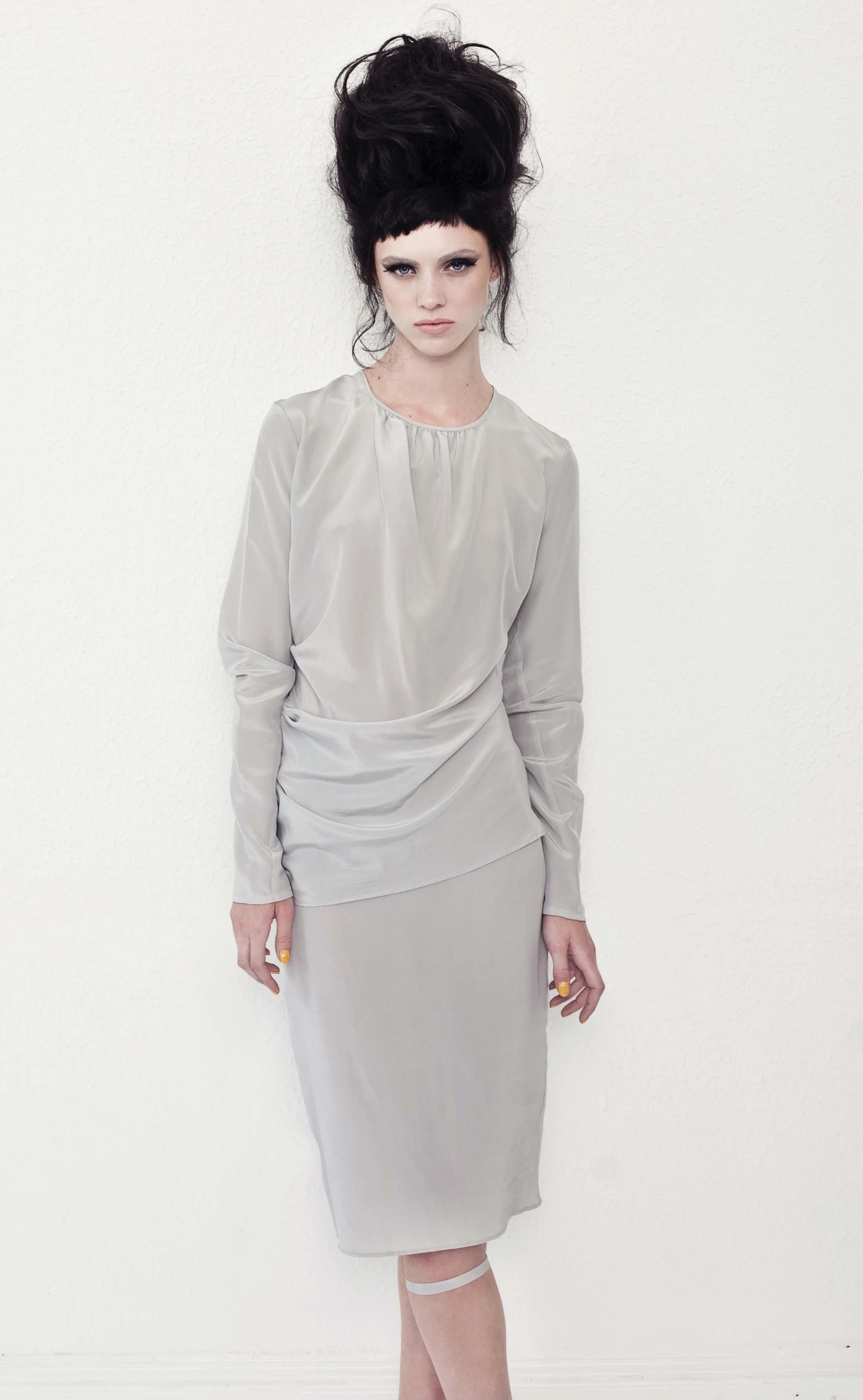 A woman with dark, voluminous hair styled in an upswept manner, wearing a light beige satin dress with long sleeves and a gathered neckline, standing against a plain white wall.