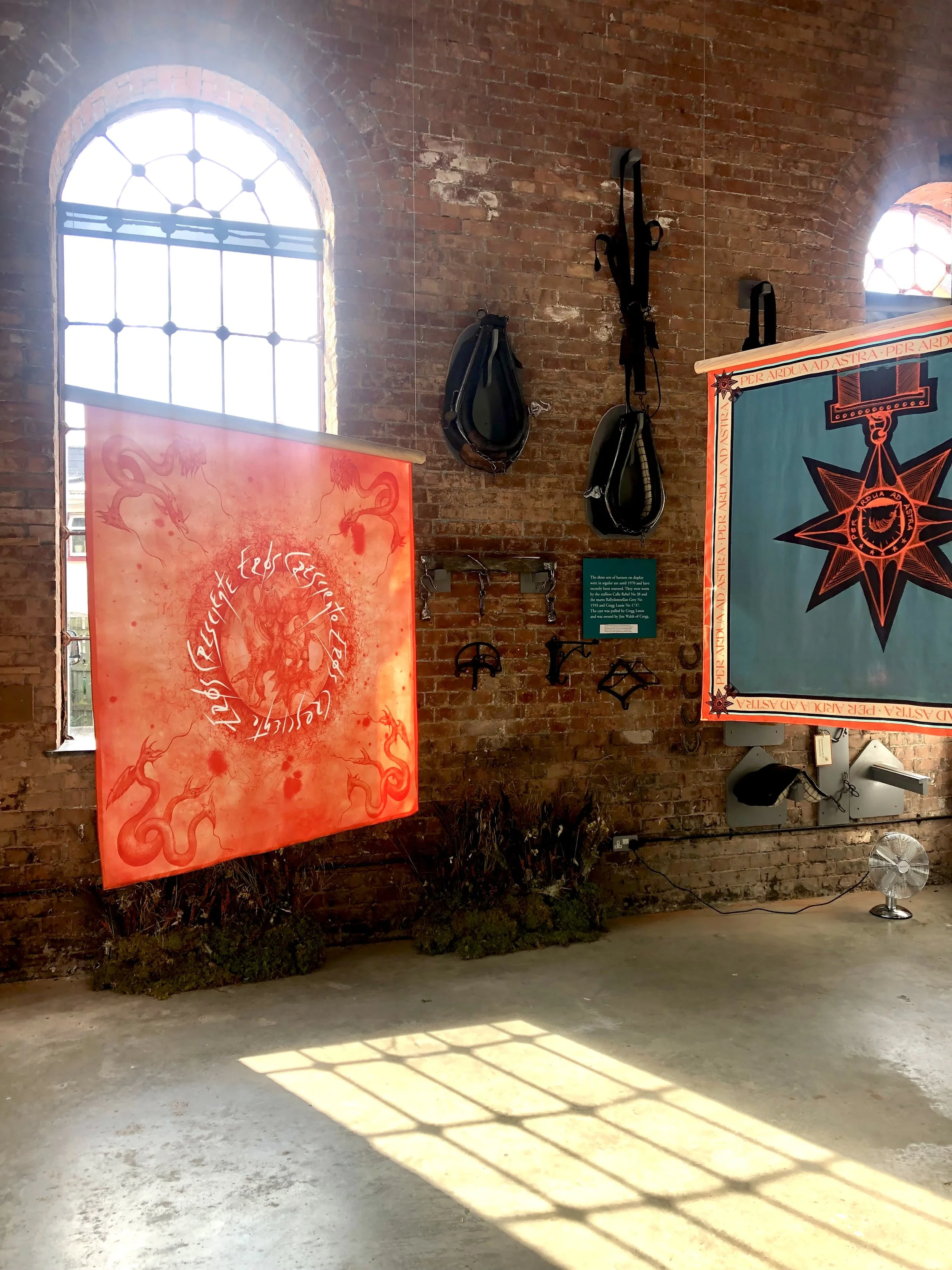 Exhibition space with brick walls, large windows, hanging banners, and some wall-mounted equipment. Sunlight creates patterns on the concrete floor.