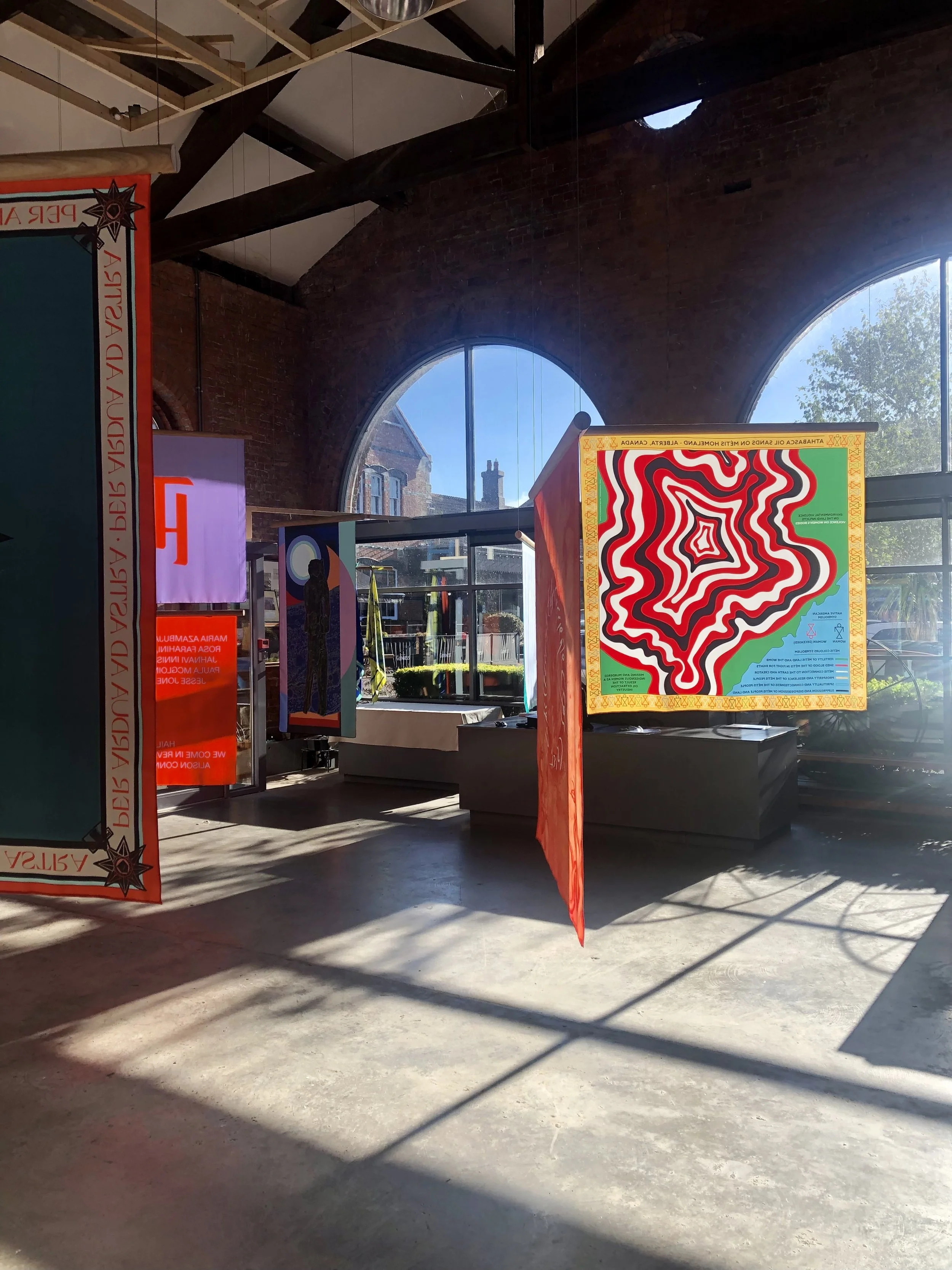 Indoor space with large windows, red and colorful artwork on display, and shadows on concrete floor.