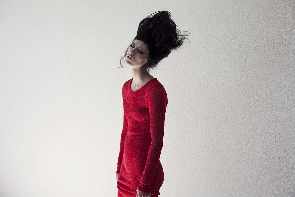 A woman with long, dark, voluminous hair wearing a tight red dress, standing against a plain, off-white textured wall, looking slightly downward.