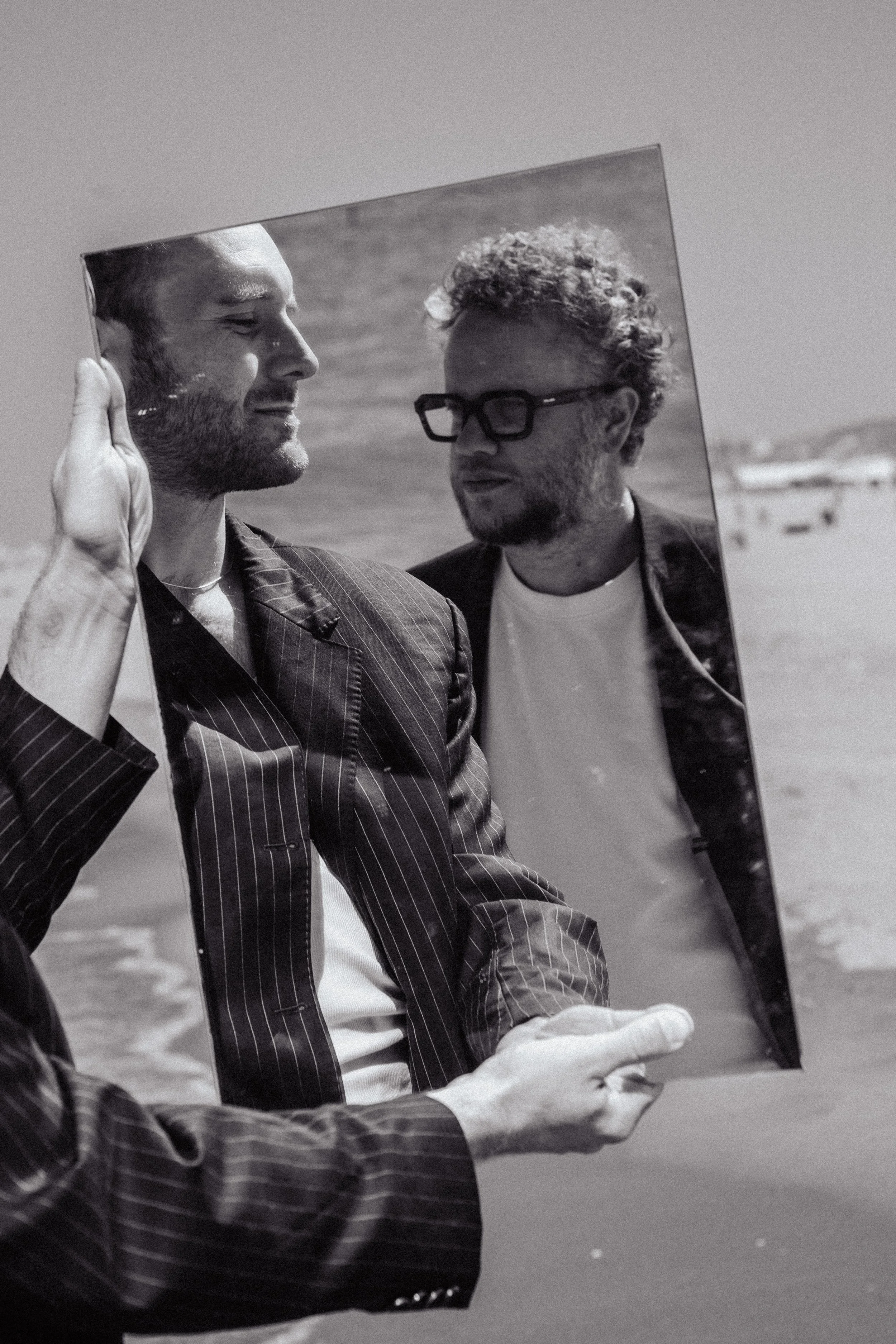 Meyendel persfoto. Een zwart-wit foto van twee mannen die een spiegel vasthouden, op het strand op een bewolkte dag.