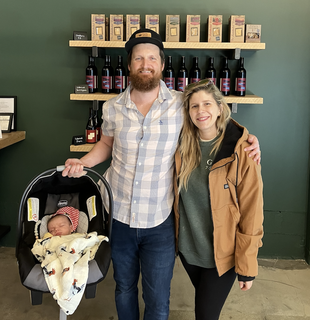 Man and woman standing in front of wine bottles holding their newborn baby