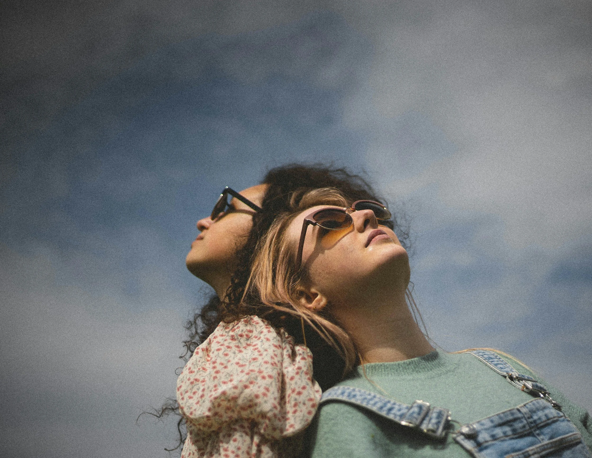 Two women wearing sunglasses looking up at the sky with clouds.