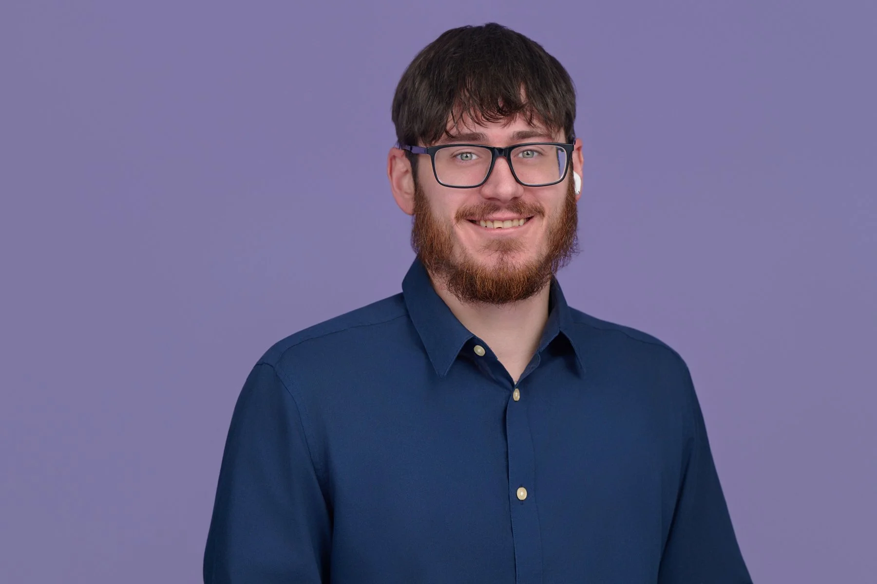 Portrait of a man with dark hair, beard, glasses, and earbud, wearing a blue button-up shirt, smiling in front of a light purple background.