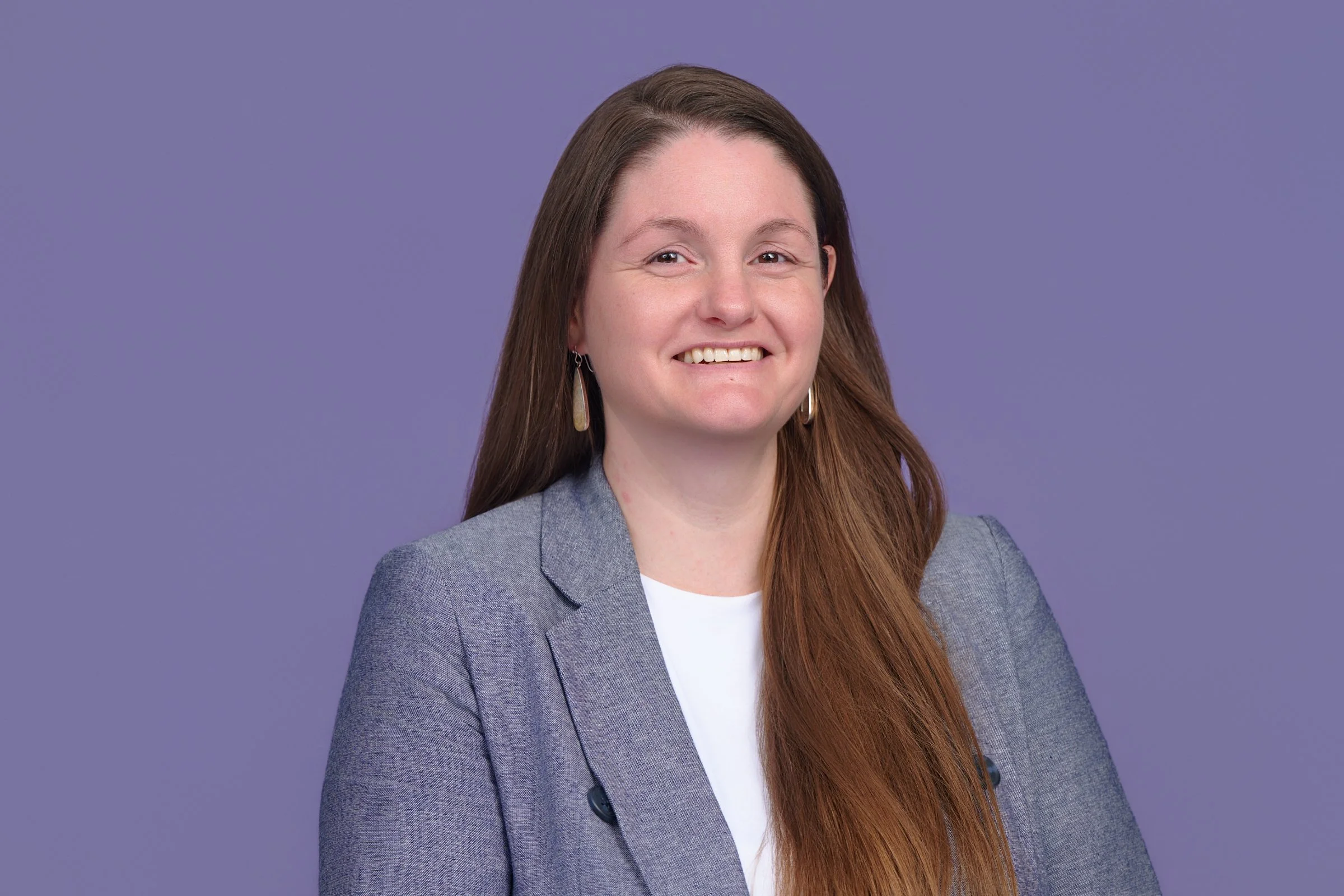 A woman with long brown hair, wearing a gray blazer and a white top, smiling and wearing earrings, against a purple background.