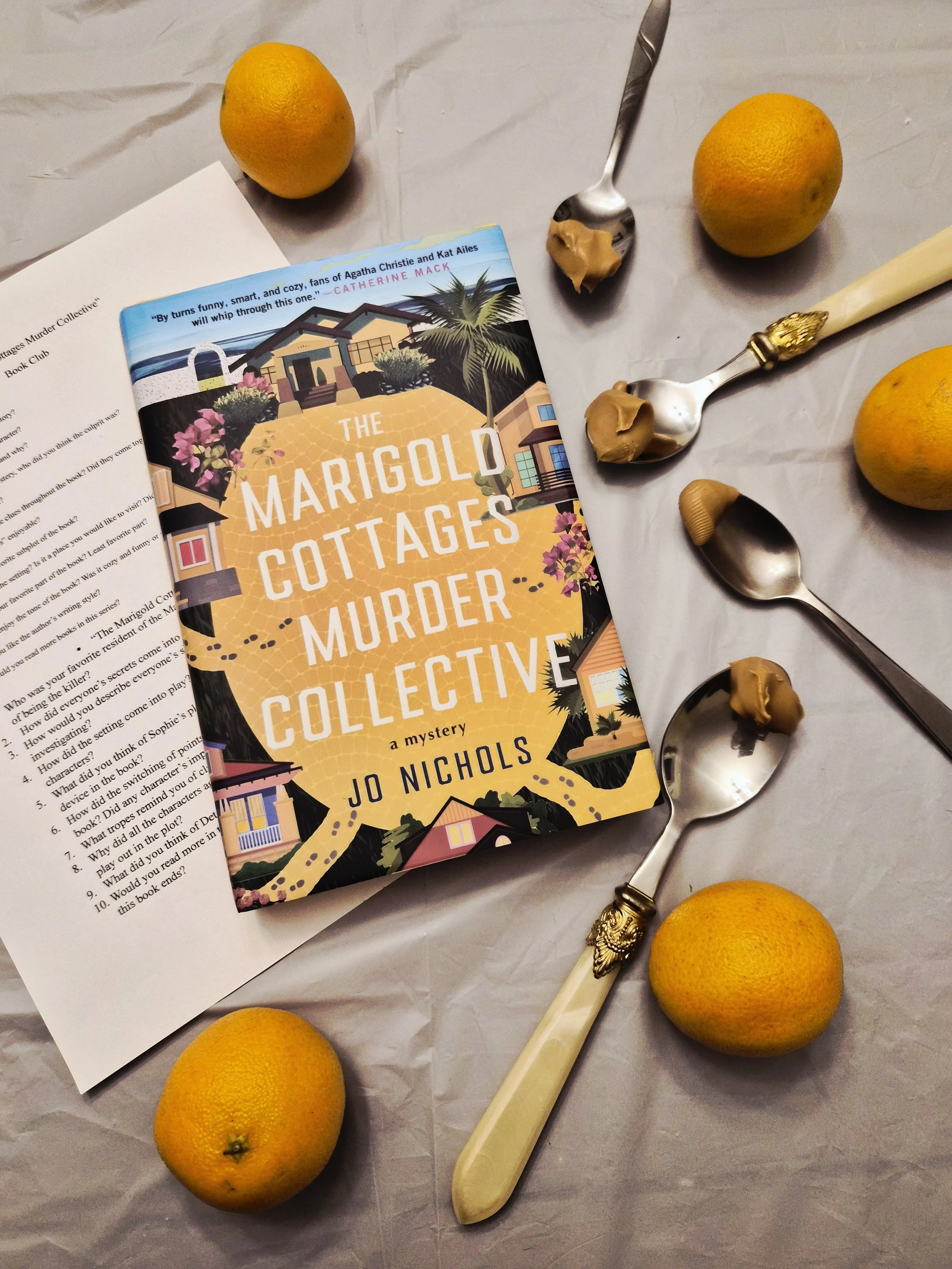 The book "The Marigold Cottages Murder Collective" surrounded by spoons of peanut butter.