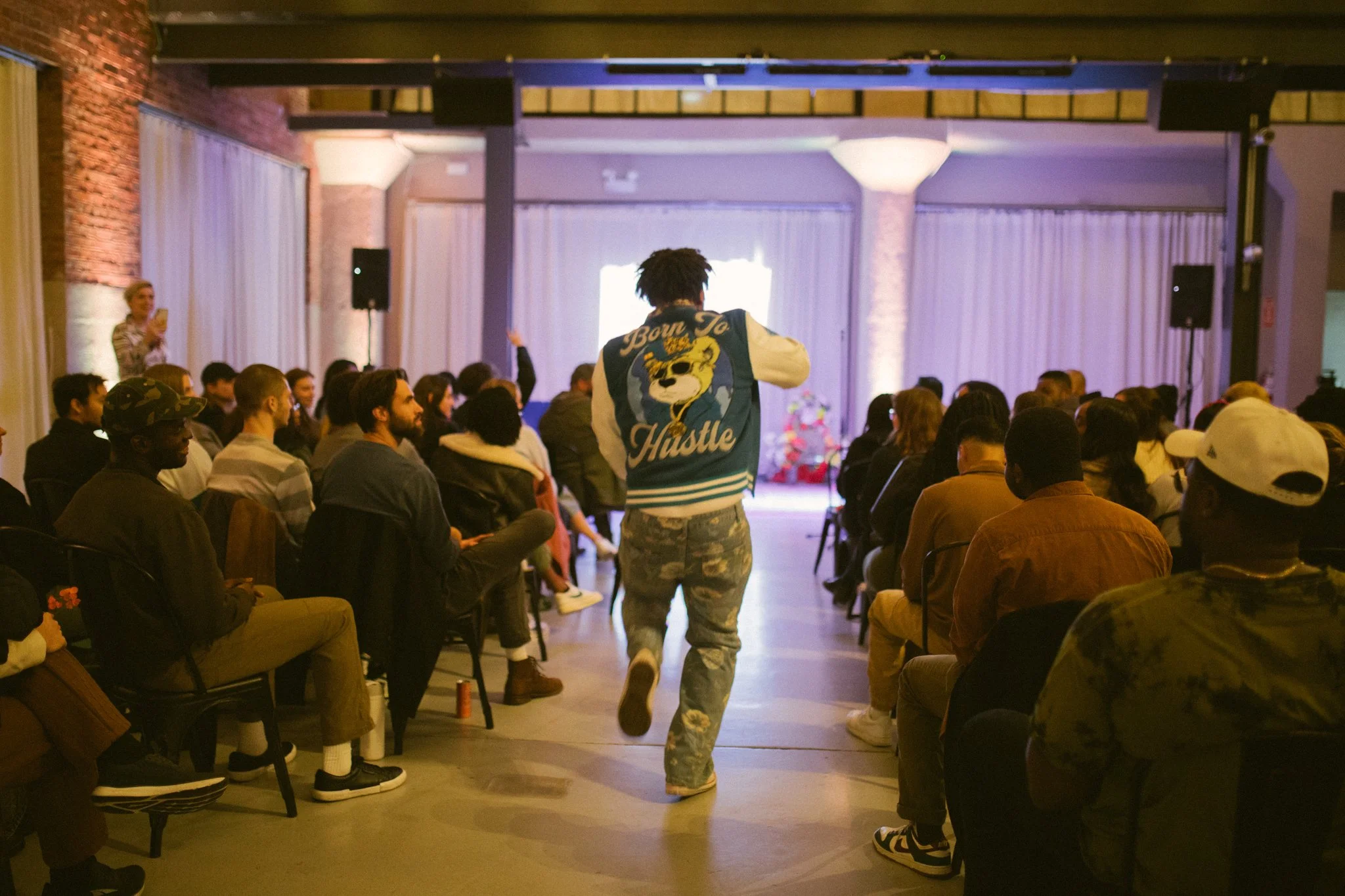 A man with dreadlocks wearing a varsity jacket and camouflage pants walking towards the back of a seated audience in a dimly lit event space with brick and curtain walls.